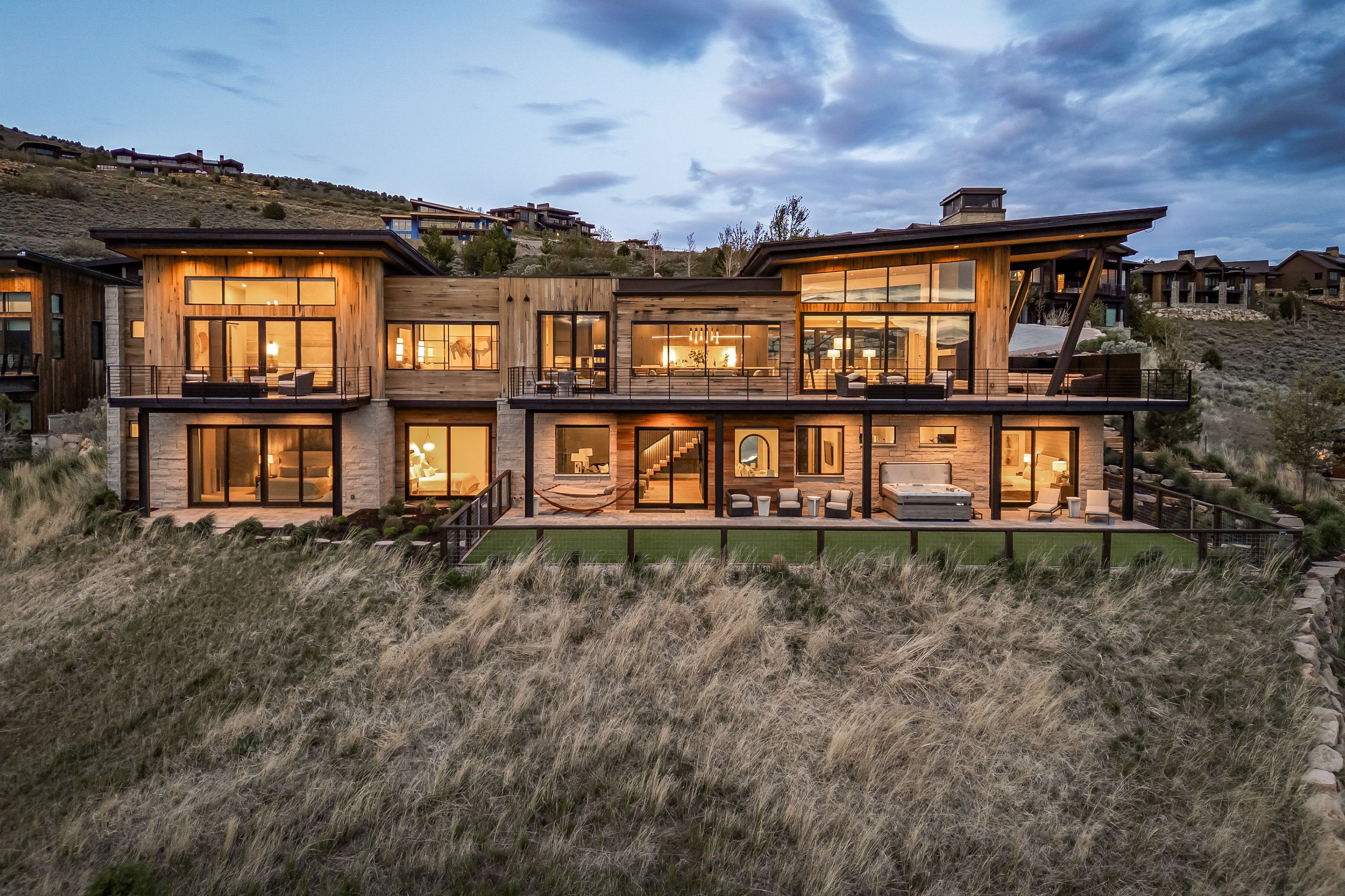 Modern architectural design featuring extensive glass window walls, multiple balconies, a lower-level patio with a built-in hot tub, and a tiered structure