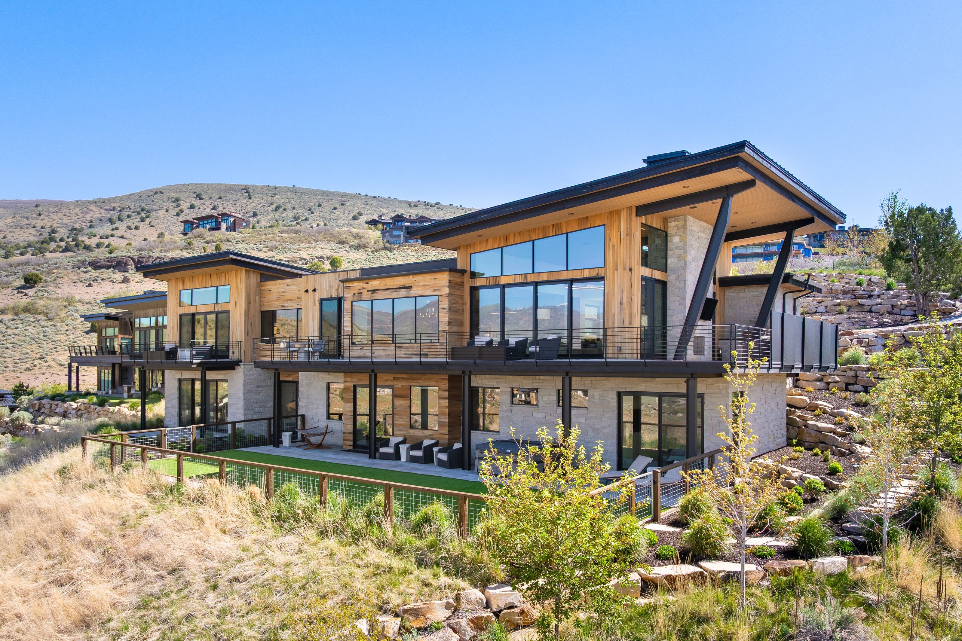 Contemporary multi-level residence featuring expansive window walls, wood-finish siding, stone-finish accents, multiple outdoor balconies with cable railings, and terraced landscaping