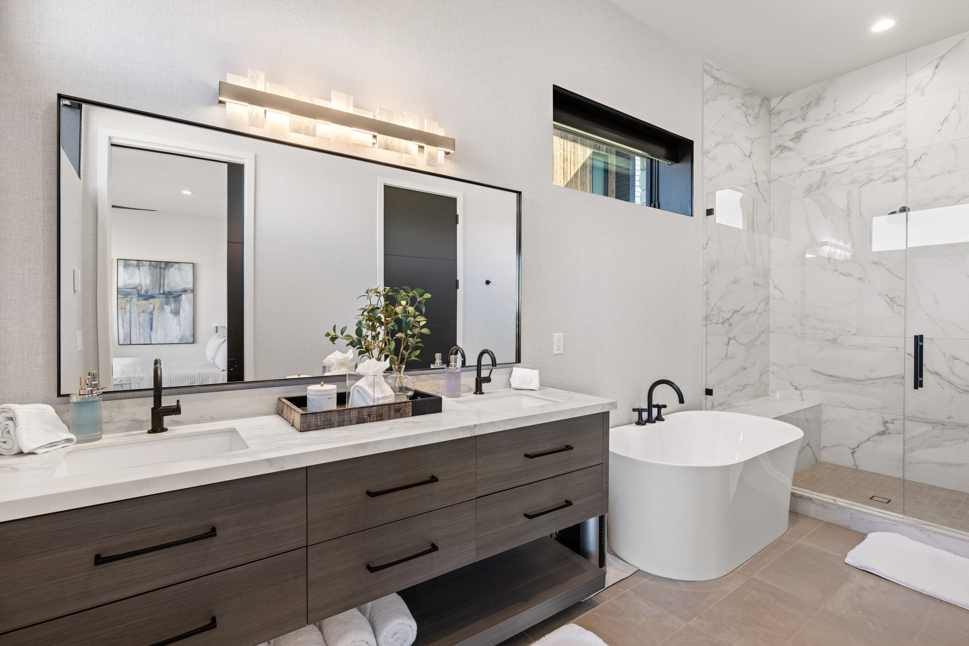 Contemporary bathroom featuring a freestanding soaking tub, a dual vanity with wood-finish cabinetry and undermount sinks, a spacious glass-enclosed shower with a bench, and marble-look wall tiling