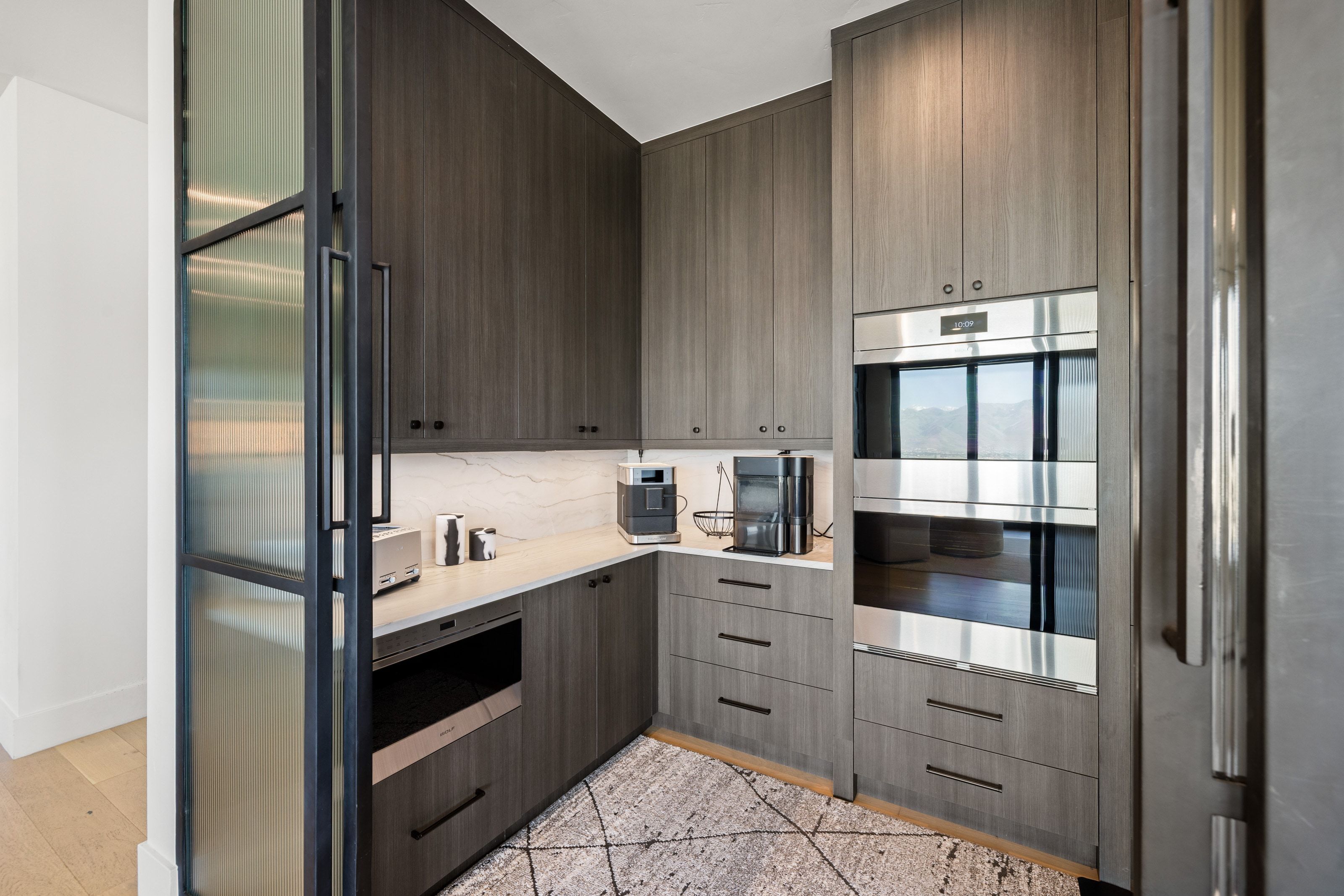 Modern kitchen area featuring dark wood-finish cabinetry, light-toned countertops with a matching backsplash, and built-in stainless steel double ovens