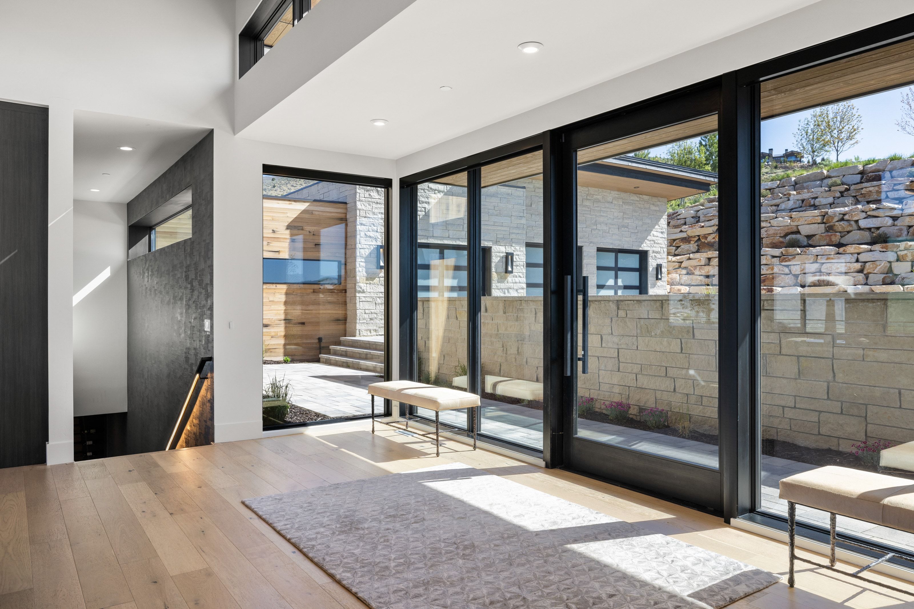 Expansive sliding glass doors with black framing, light wood-finish flooring, recessed lighting, a stone retaining wall, and a wood-paneled exterior entryway