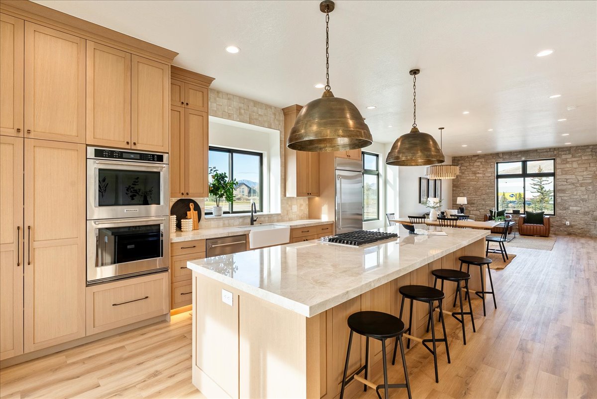 Expansive kitchen featuring light wood-finish cabinetry, built-in double ovens, a gas range on a substantial island with seating, pendant lighting, and wood-finish flooring