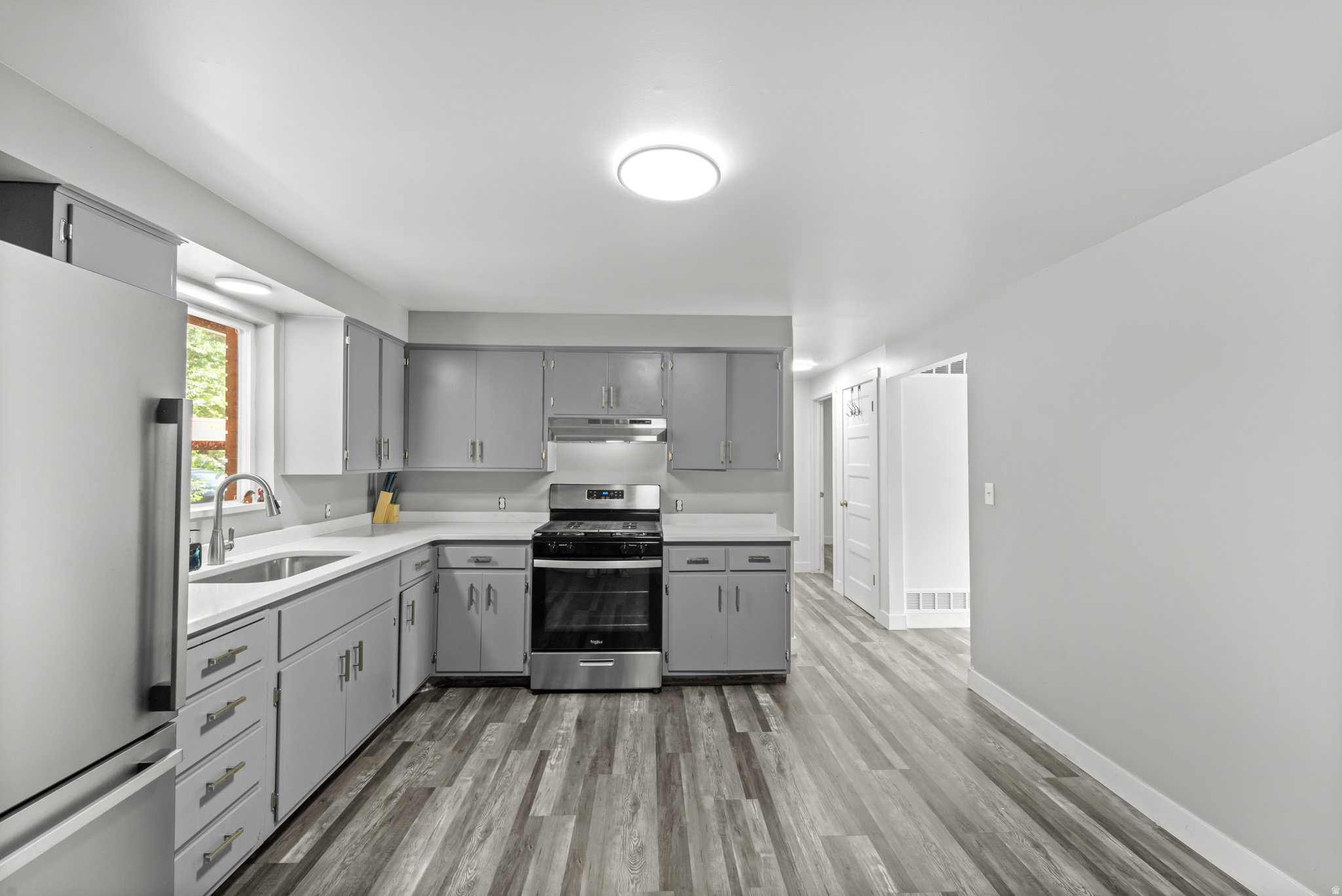 Kitchen featuring wood-finish flooring, light grey cabinetry with silver hardware, stainless steel appliances, a white sink with a gooseneck faucet, and white countertops