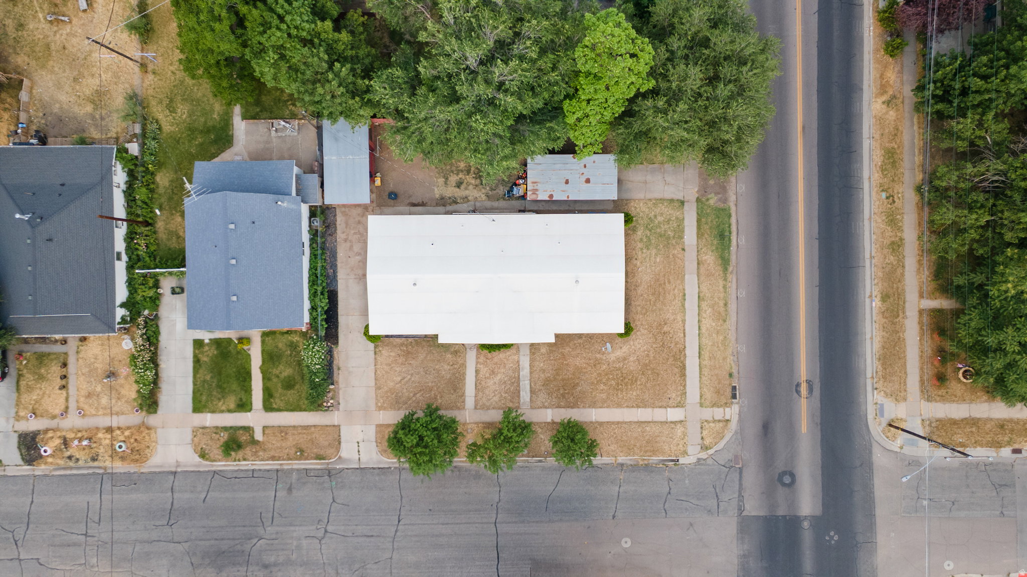 Aerial perspective showcasing a property with a light-colored roof, surrounded by mature trees and a paved street