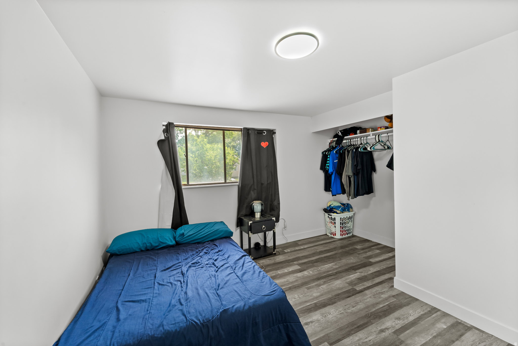 Bedroom featuring wood-finish flooring, a window with black curtains, an integrated closet with shelving and hanging rod, a ceiling-mounted light fixture, and white walls