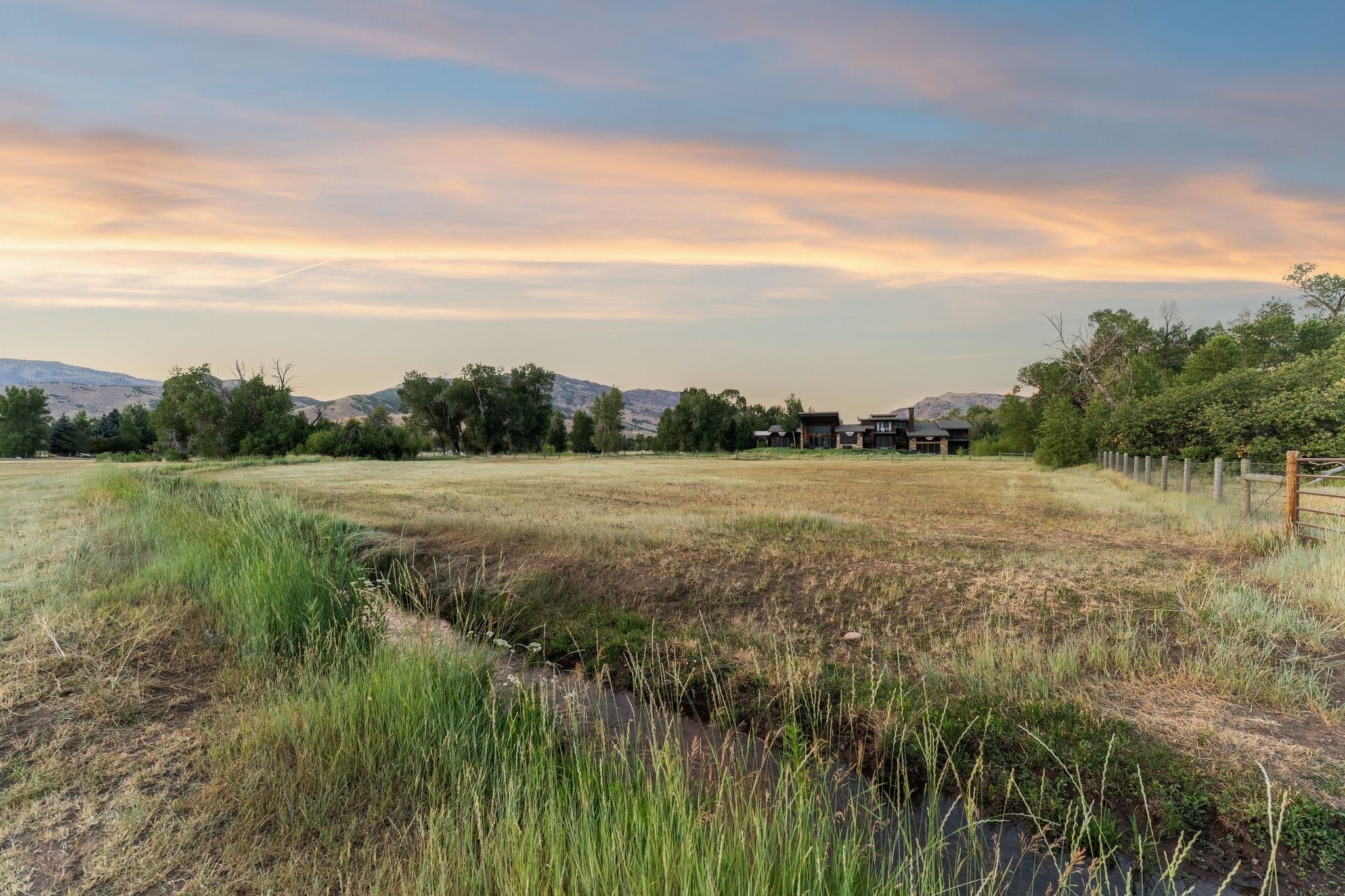Expansive natural landscape featuring a rustic fence line and distant structures, complemented by a serene stream and varied foliage