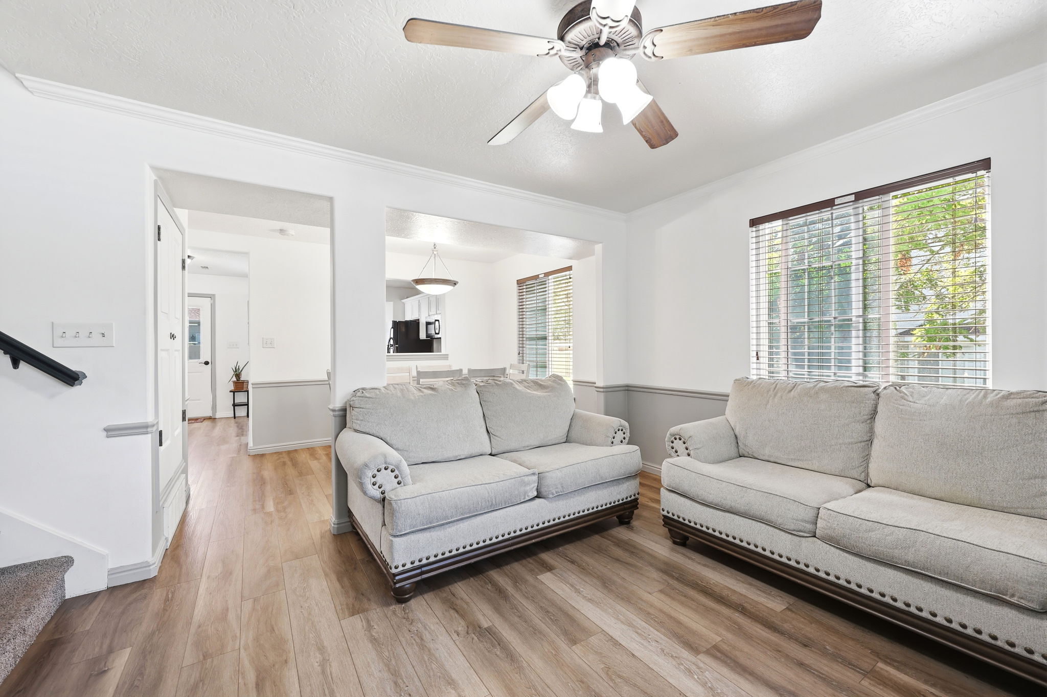 Living area featuring wood-finish flooring, a ceiling fan with integrated lighting, and a large window with blinds