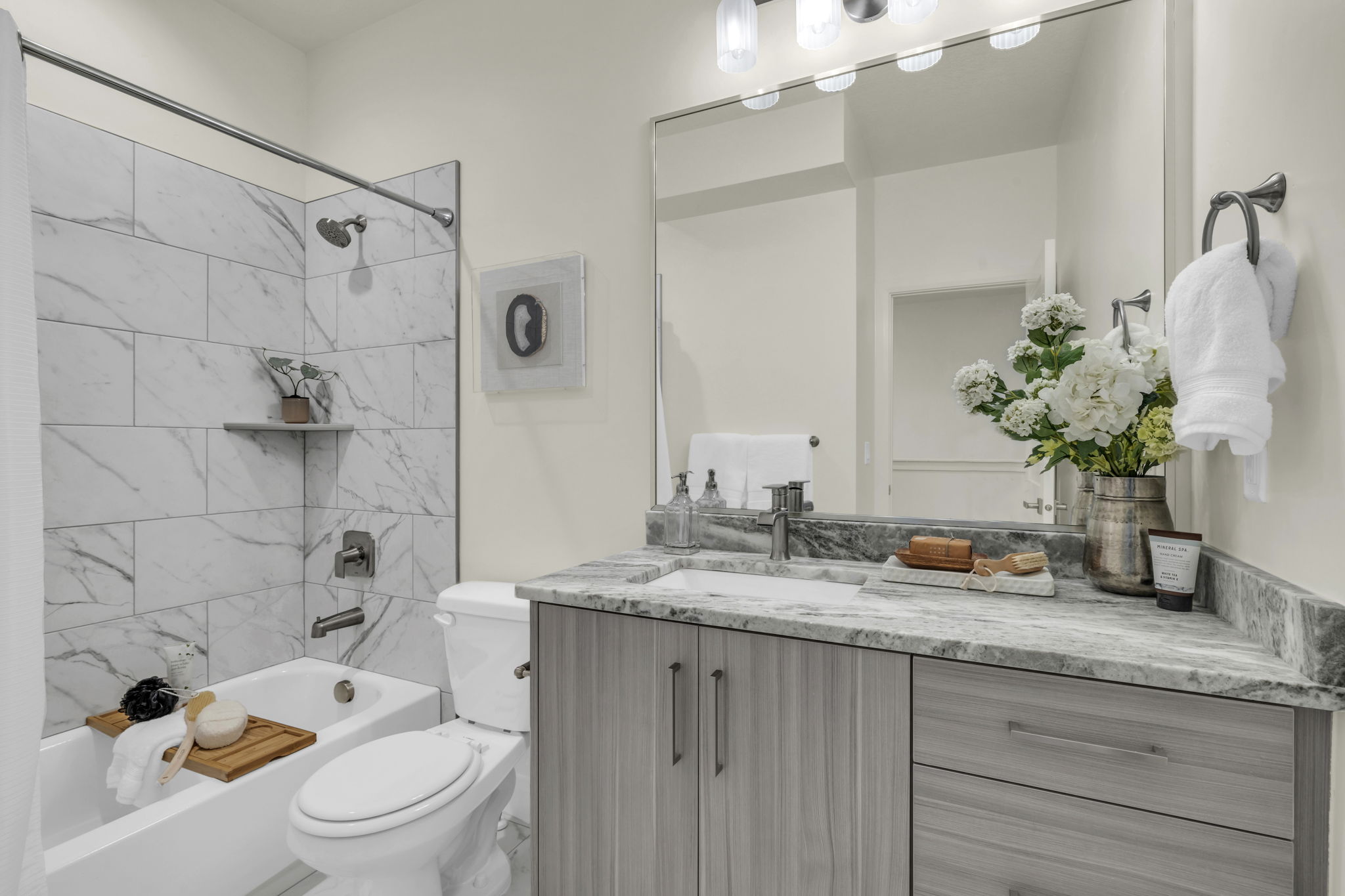 Bathroom featuring a white subway tile tub surround, built-in corner shelf, light wood-finish vanity with a single basin, gray countertop, and an expansive wall mirror