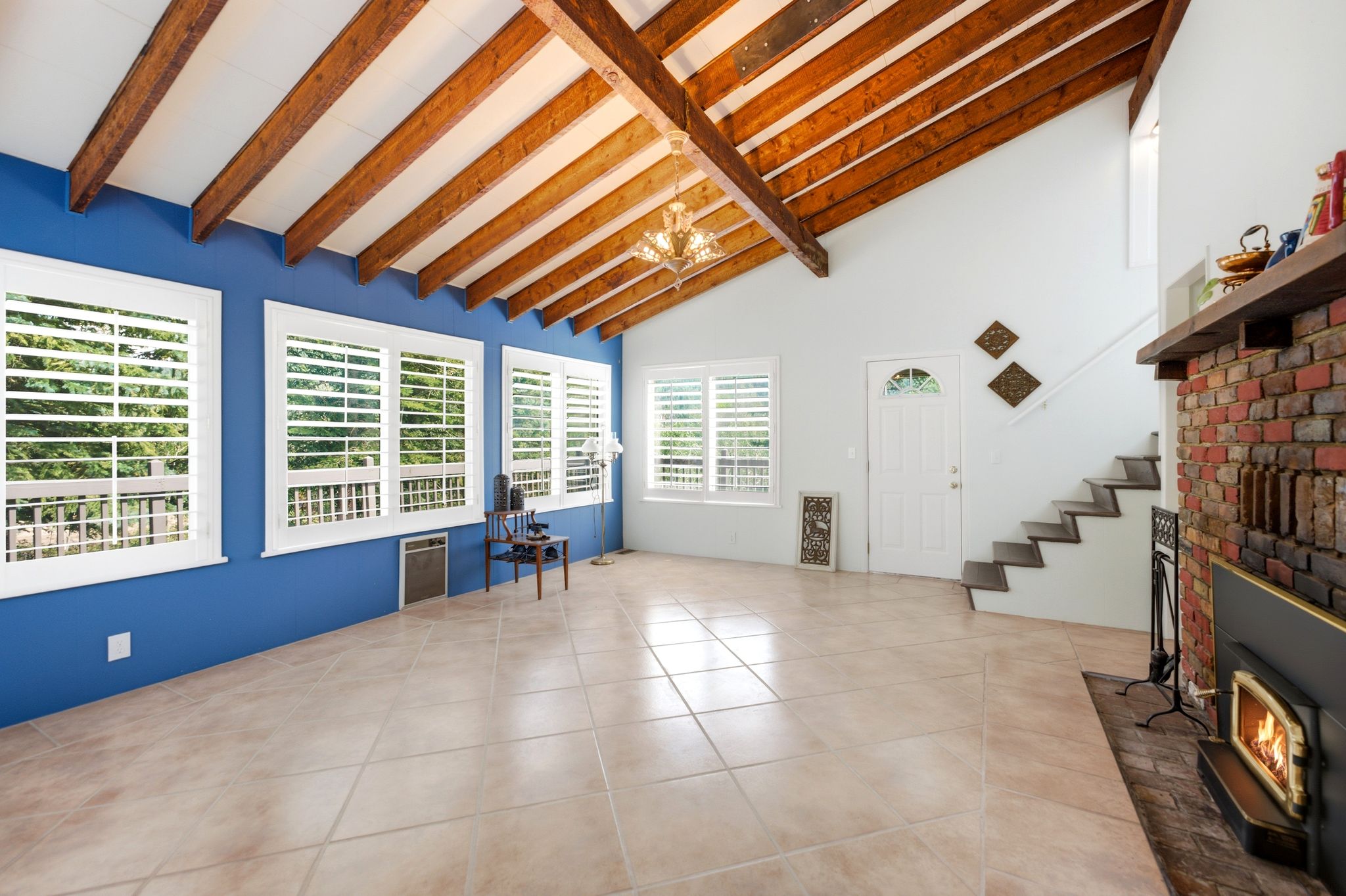 Vaulted ceiling with exposed wood beams, tile flooring, multiple windows with plantation shutters, brick fireplace with wood mantel, and an interior staircase