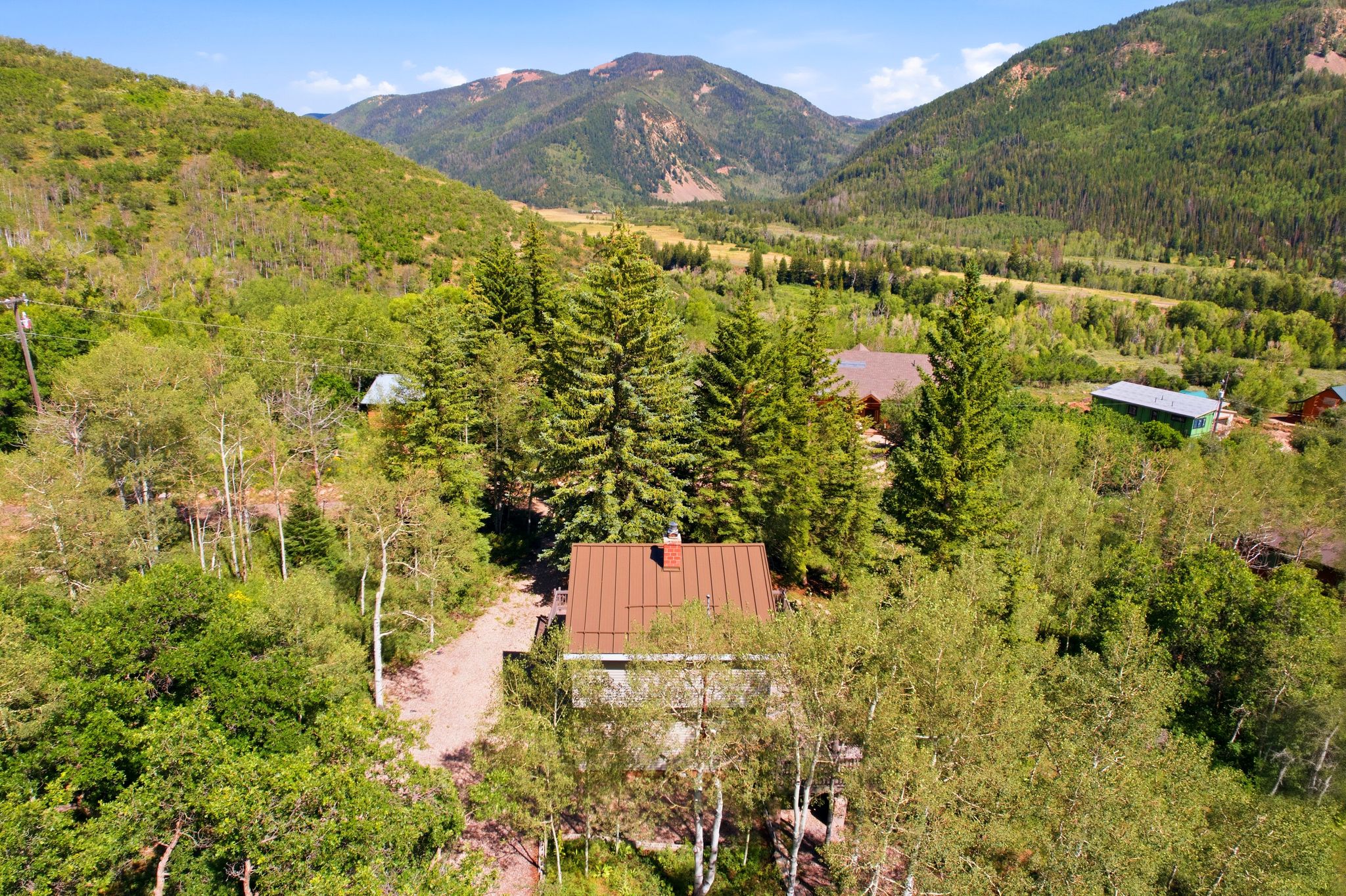 Mountainous landscape featuring a dwelling with a brown pitched roof, surrounded by numerous mature trees and a gravel pathway