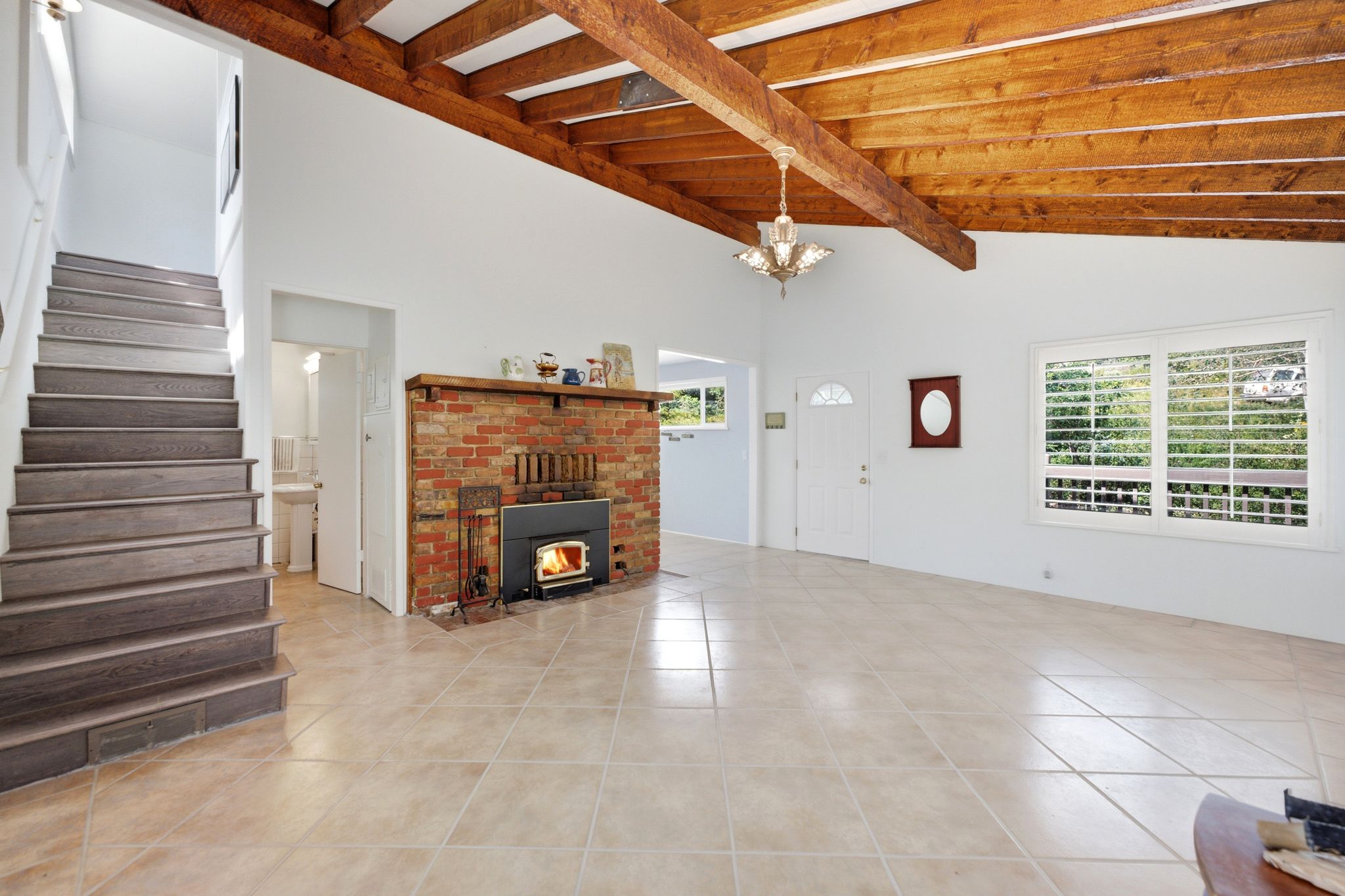 Spacious living area featuring a vaulted ceiling with exposed wood beams, a brick fireplace with a wood-burning insert, an open-tread staircase with wood-finish risers, large windows with interior shutters, and neutral-toned tile flooring