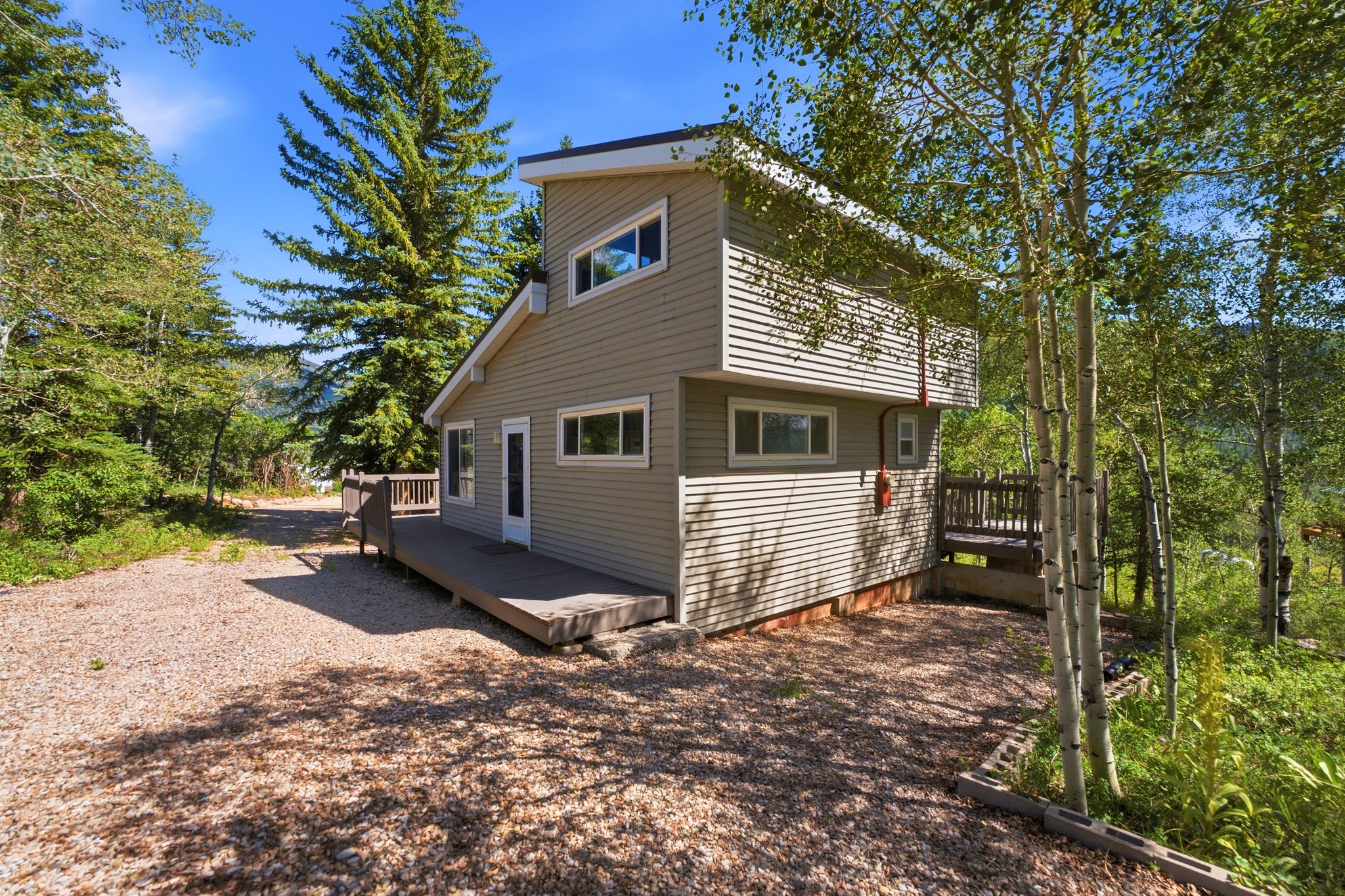 Two-story home with horizontal siding and multiple windows, featuring a gravel driveway and surrounding mature trees