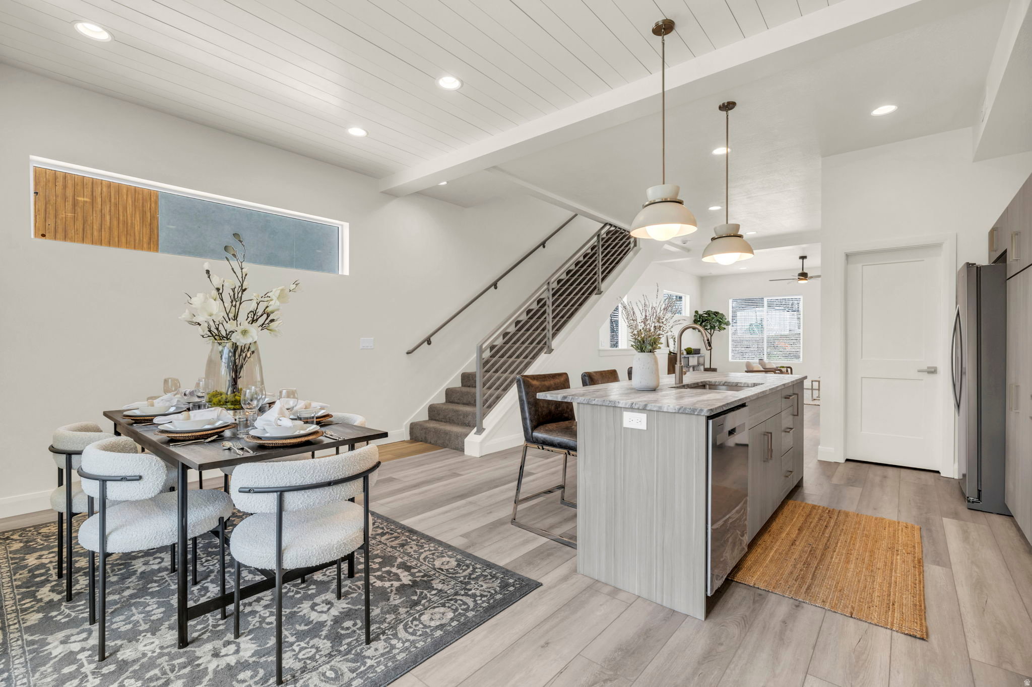 Open-concept living space with wood-finish flooring, a contemporary kitchen island with a stone countertop, modern pendant lighting, and a staircase with metal railings