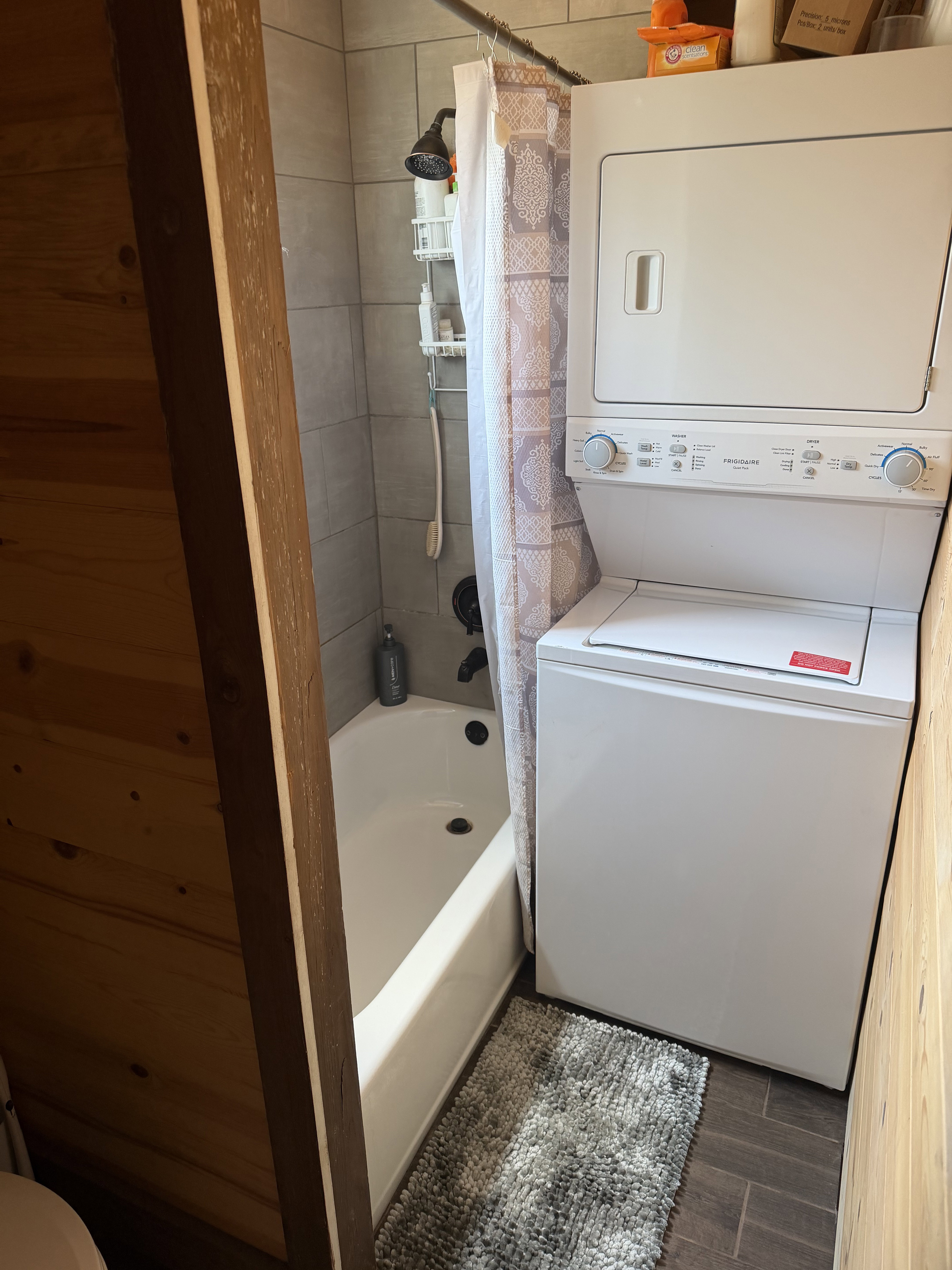 Vertical stacked washer and dryer unit beside a white bathtub with a tiled shower surround and dark bronze fixtures