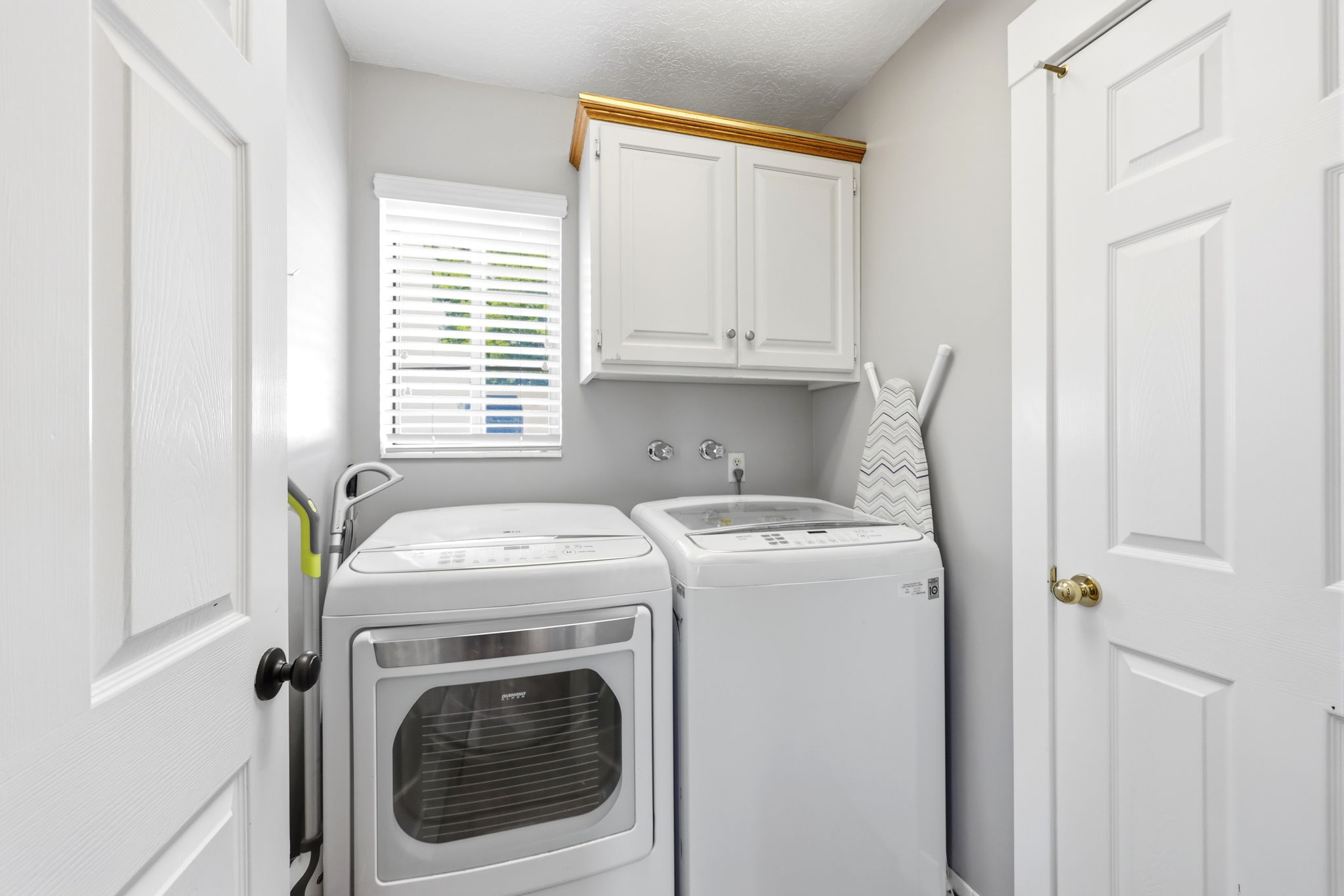Dedicated laundry area featuring white shaker-style cabinetry with gold trim, a window with horizontal blinds, and paneled doors with black and brass hardware