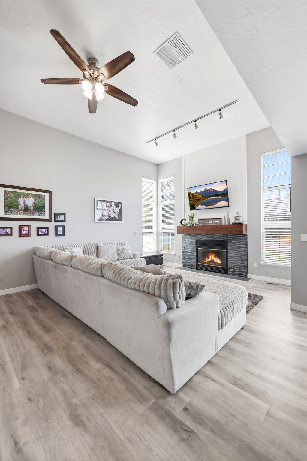 Living area featuring high ceilings, wood-finish flooring, a stone-clad fireplace with wood mantel, a ceiling fan with integrated lighting, and multiple windows