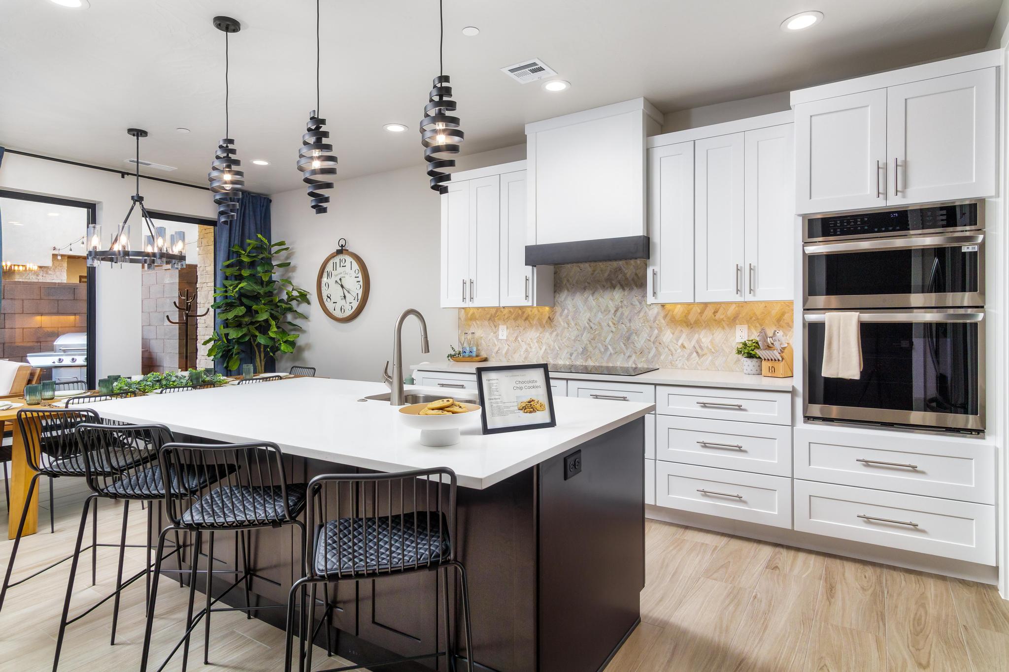 Spacious kitchen featuring a large central island with seating, white shaker cabinetry, stainless steel double ovens, a decorative range hood, and light wood-finish flooring