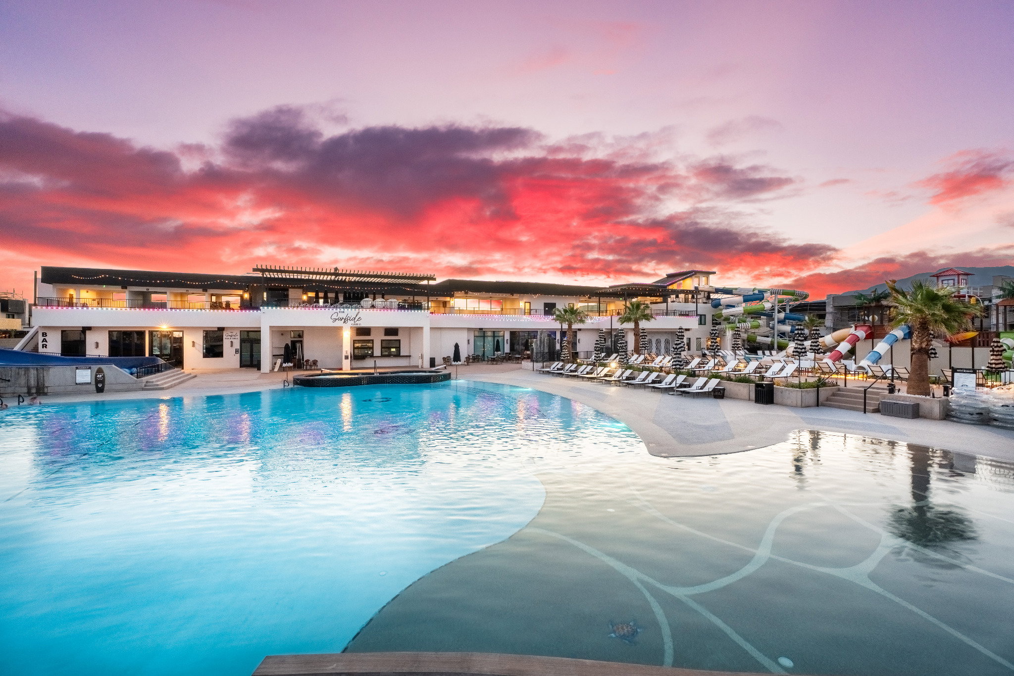 Expansive aquatic area featuring a large swimming pool with integrated patterns, a multi-level building with outdoor lounging spaces, a dedicated water park area with slides, and mature palm trees