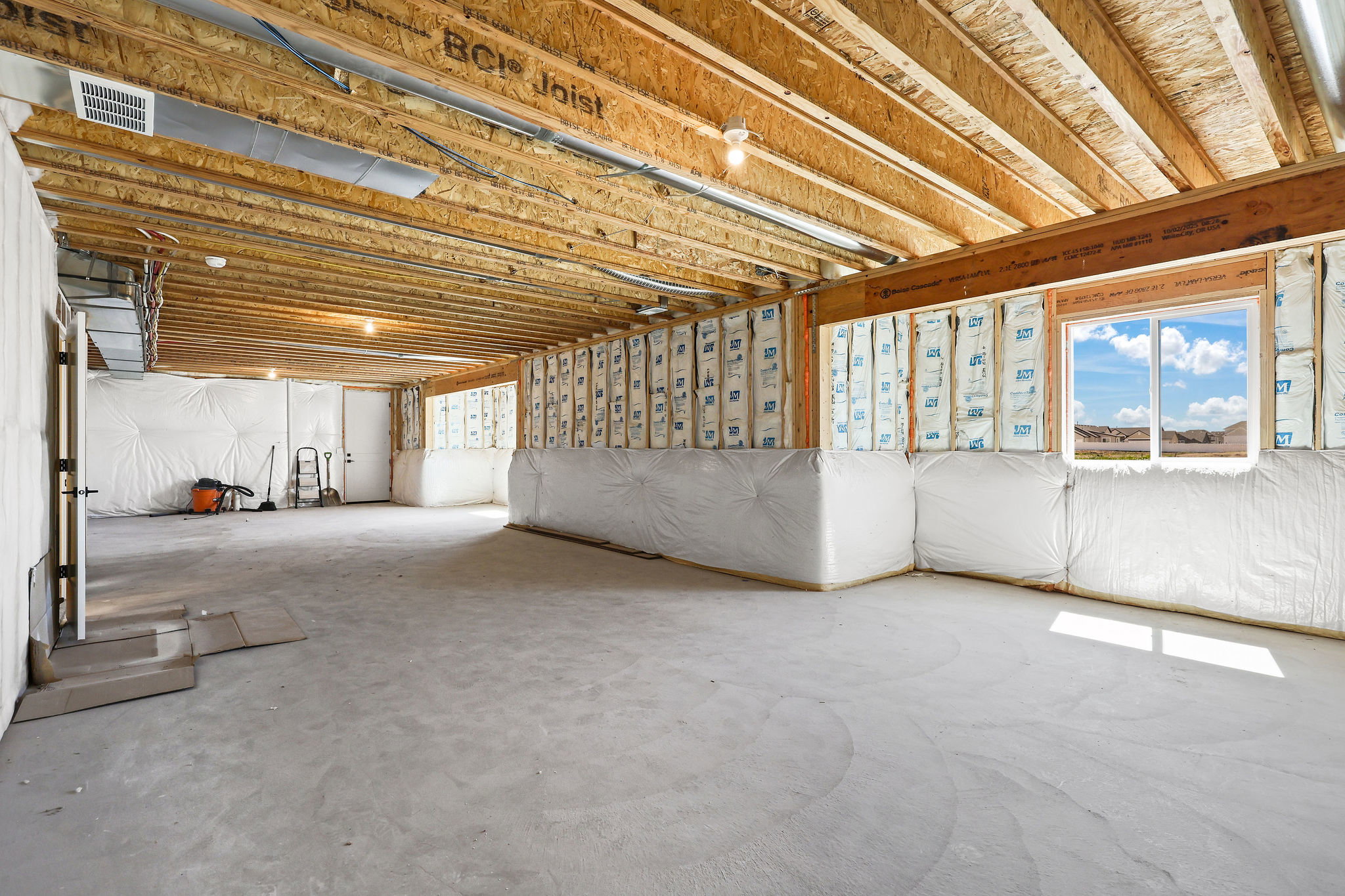 Unfinished basement space with exposed wood joists and subfloor, featuring a large window with exterior views