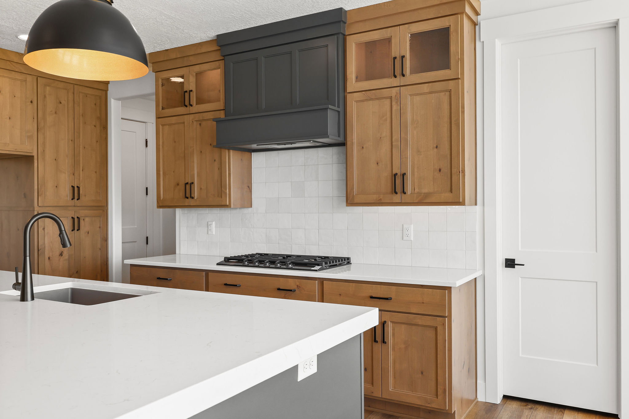 Kitchen featuring light wood cabinetry, a dark gray custom range hood, white subway tile backsplash, white countertops, and a kitchen island with an integrated sink and black faucet