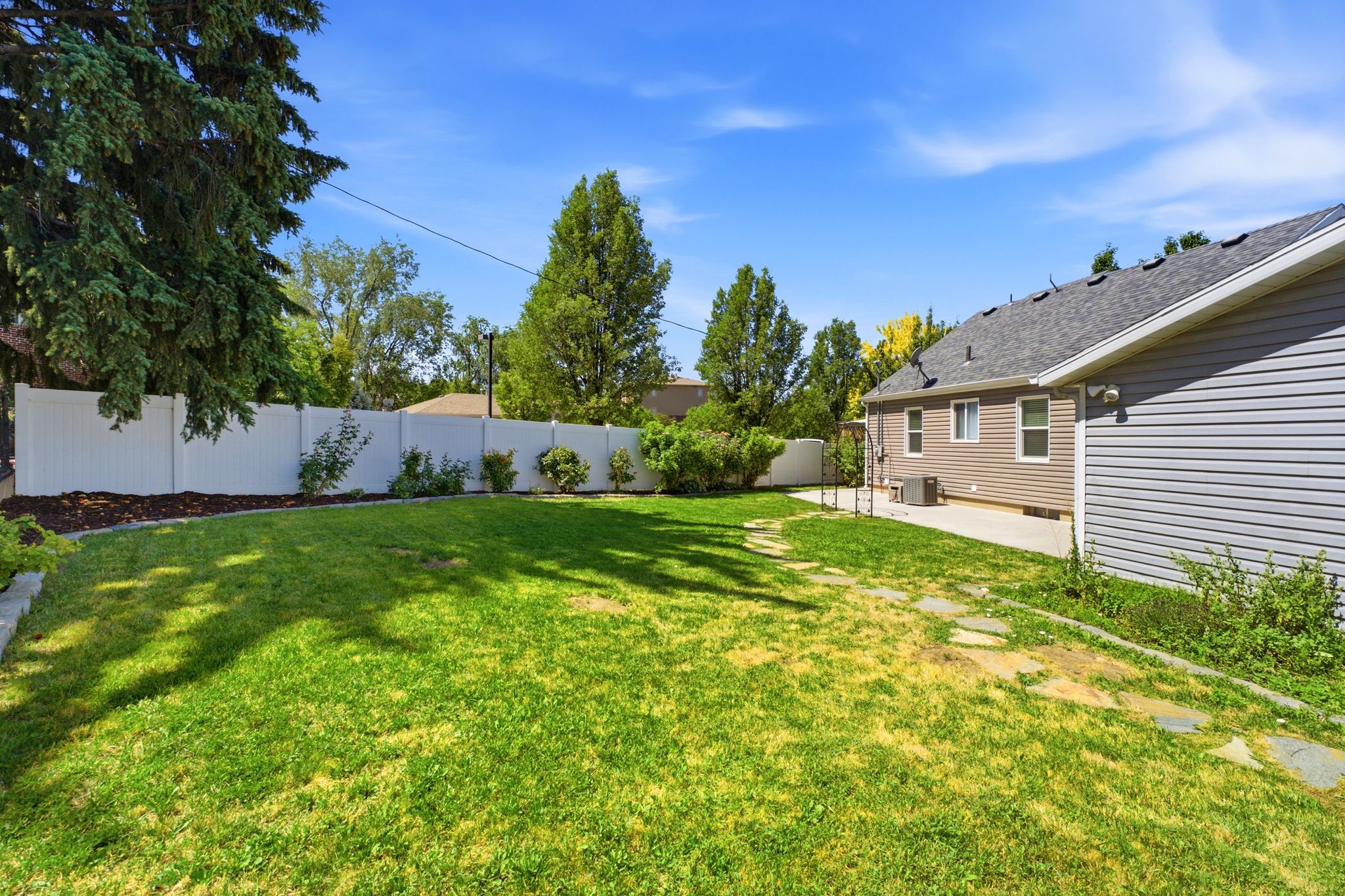 Expansive lawn area featuring a white privacy fence, established trees, a concrete patio, and a stone-paver walkway