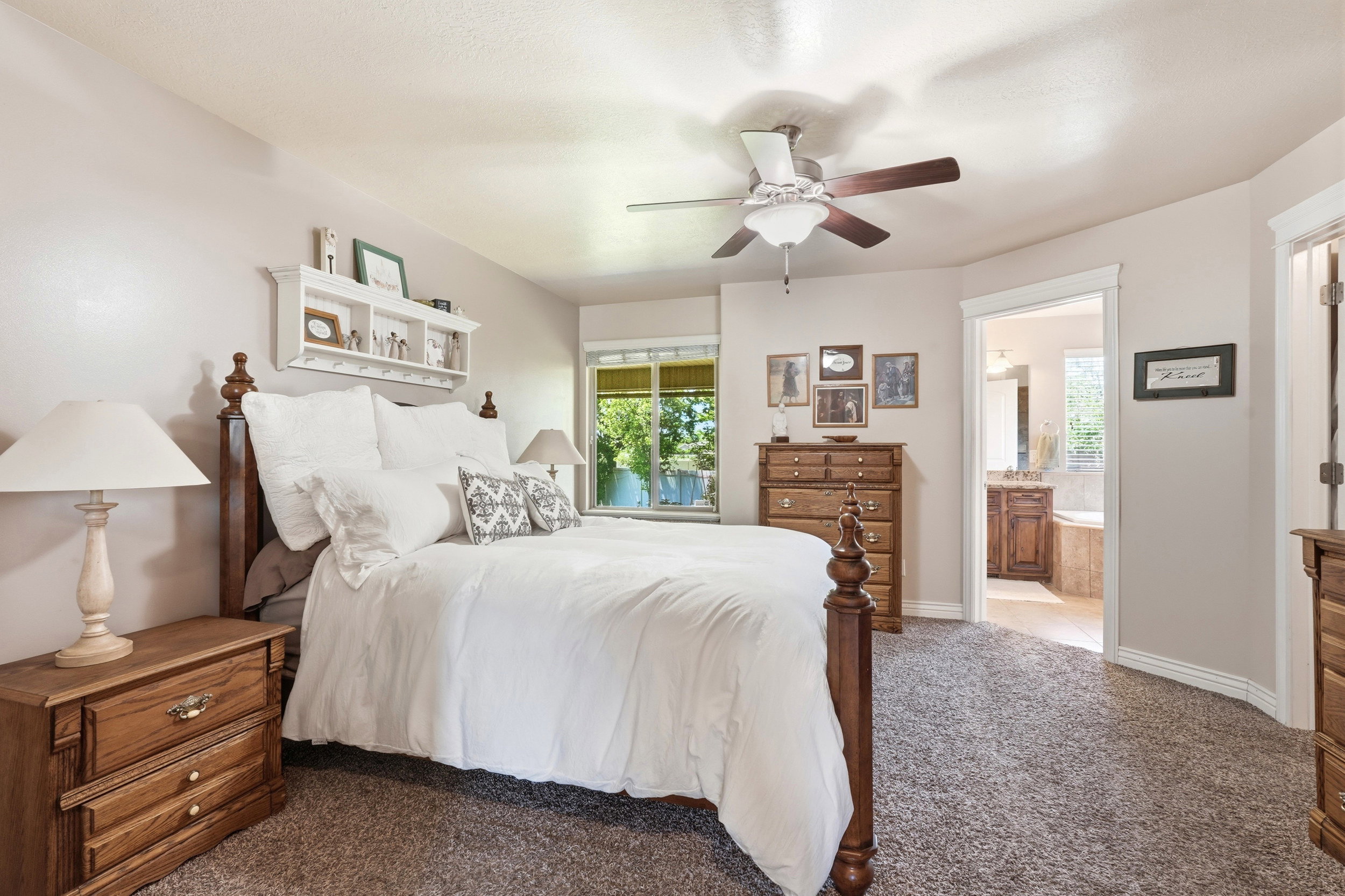 Carpeted bedroom featuring a ceiling fan with light fixture, natural light from exterior window, and an open doorway to a bathroom with a built-in tub