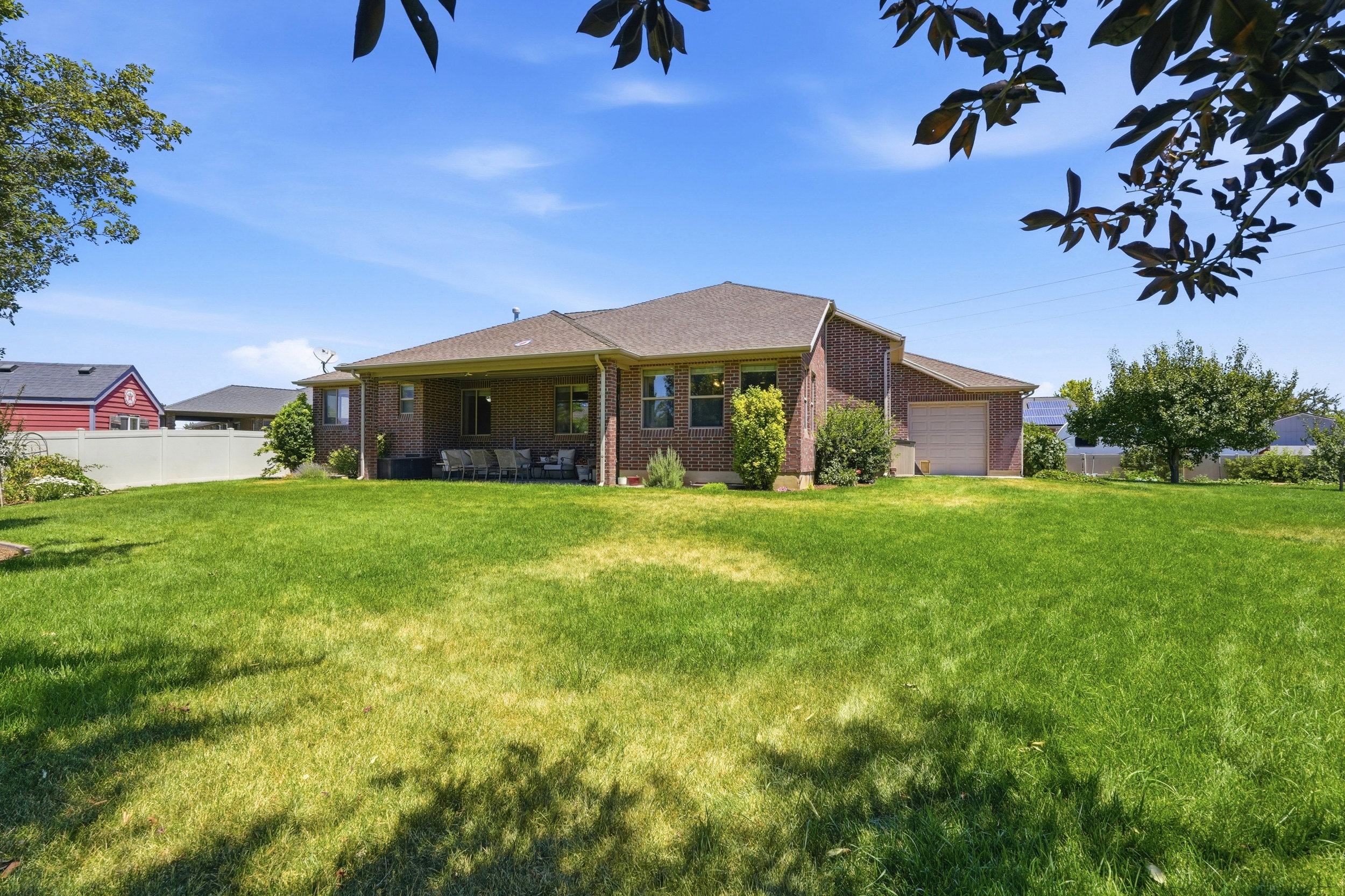 Expansive green lawn surrounds a brick residence featuring a covered patio, multiple windows with dark trim, and a detached garage with an overhead door