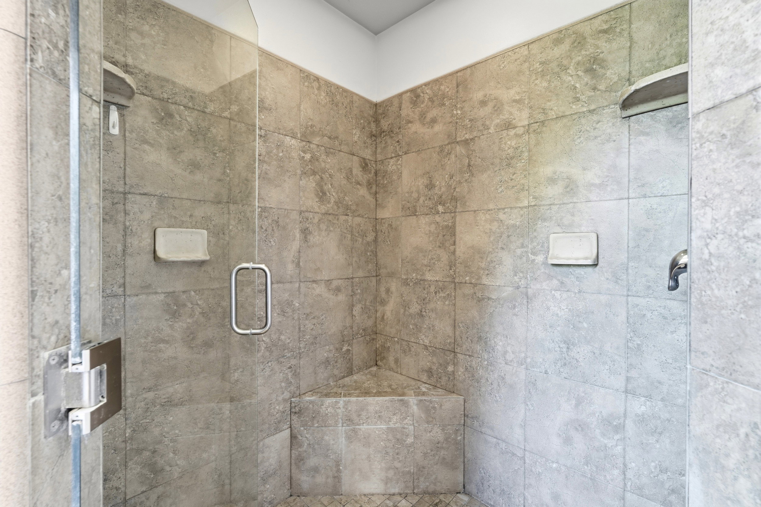 Spacious shower with stone-finish tile walls, built-in corner bench, and a glass enclosure with a polished chrome handle