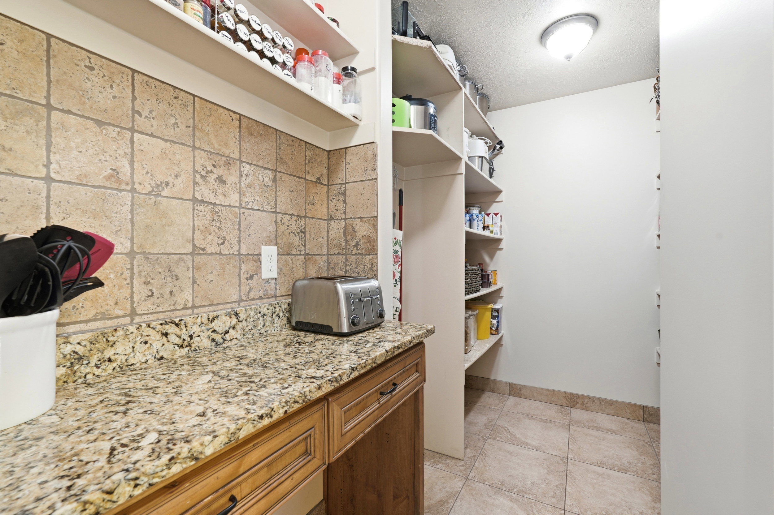 Kitchen pantry with granite-finish countertop, tiled backsplash, and wood-finish cabinetry
