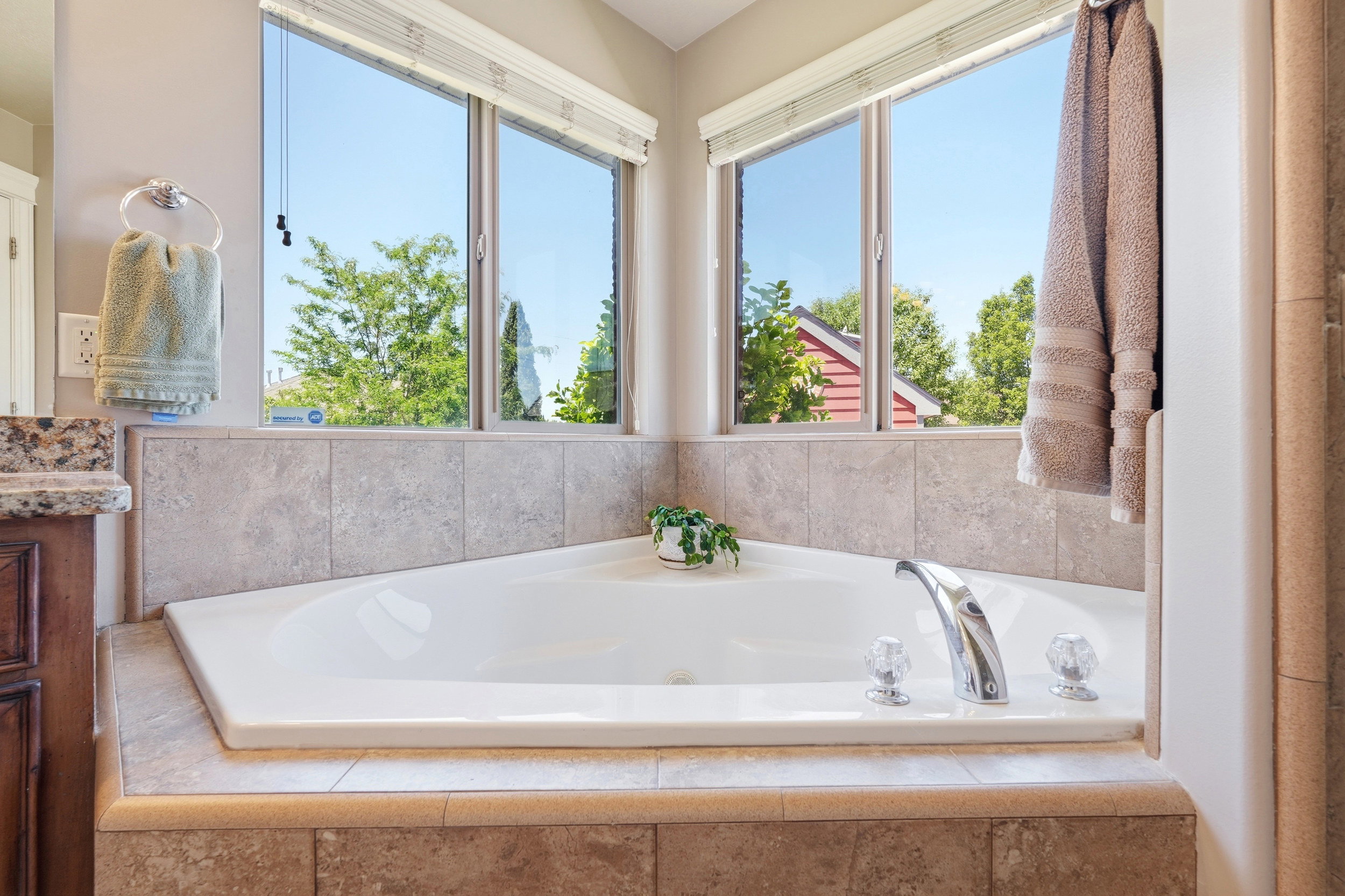 Spacious corner soaking tub featuring a chrome faucet with clear knobs, surrounded by large format tile, and framed by two corner windows with blinds