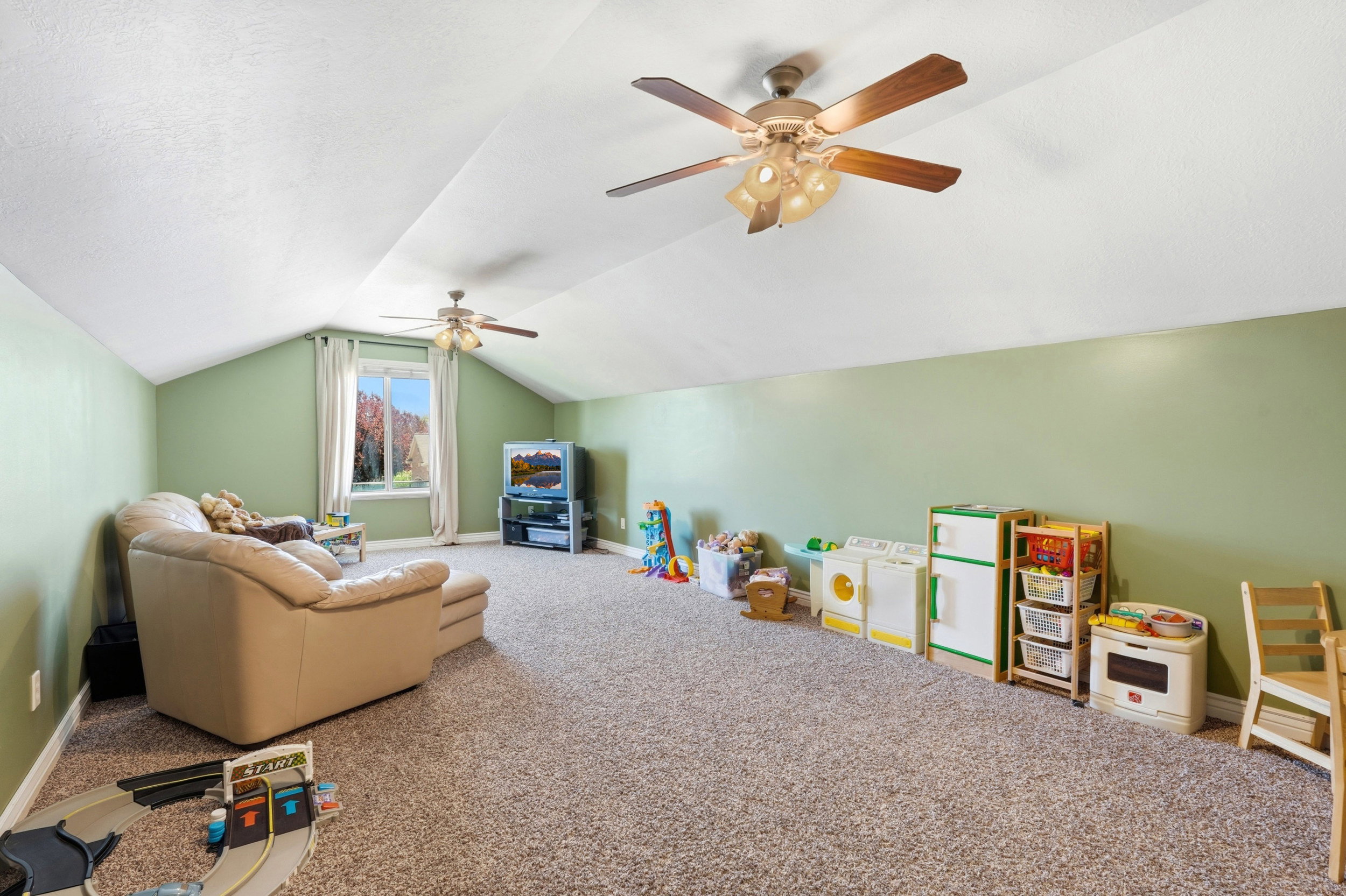 Spacious room featuring vaulted ceilings, two ceiling fans with integrated lighting, a large window, and wall-to-wall carpeting