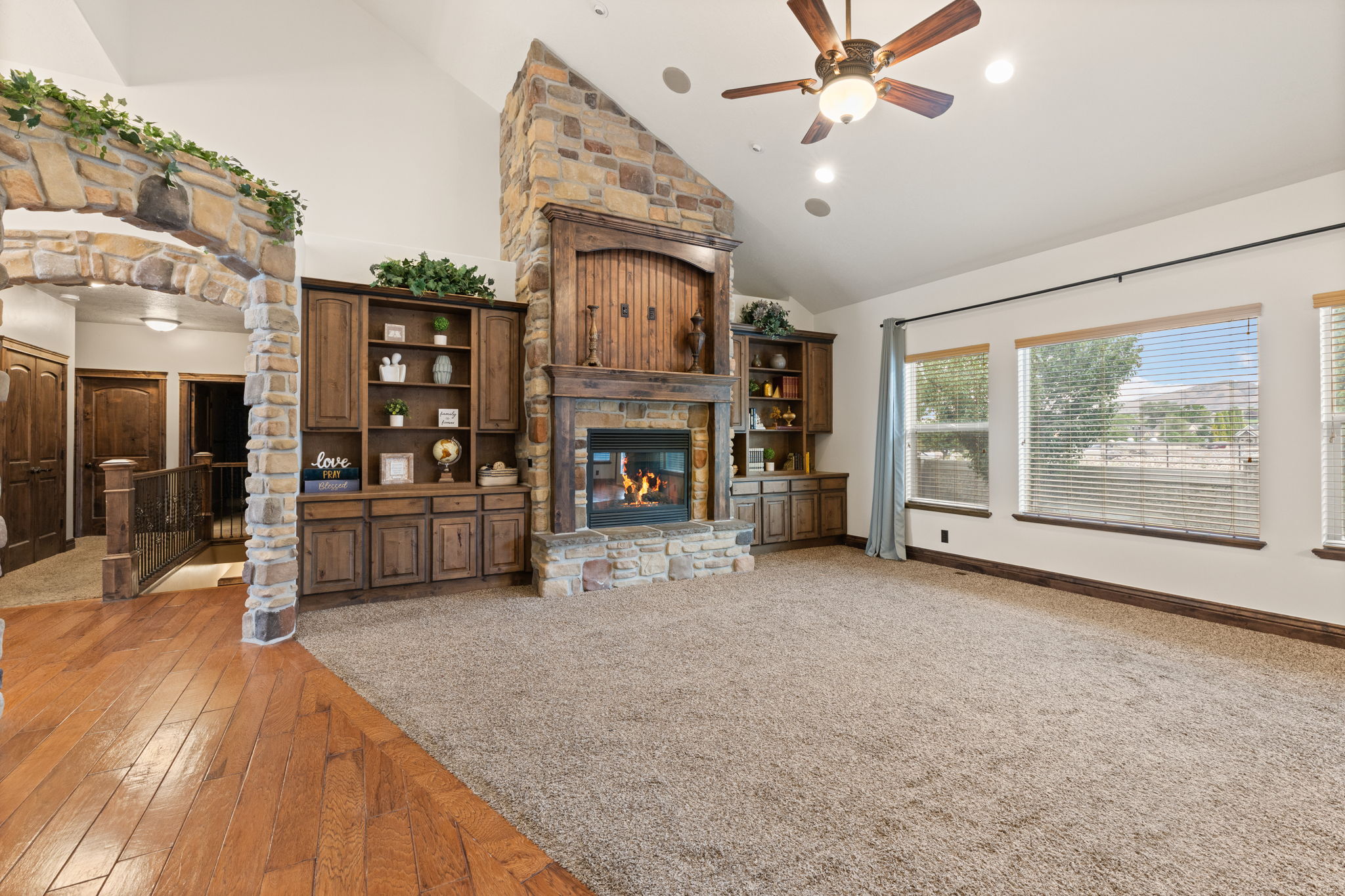 Spacious living area featuring a stone fireplace, vaulted ceilings, a ceiling fan, built-in cabinetry, and large windows