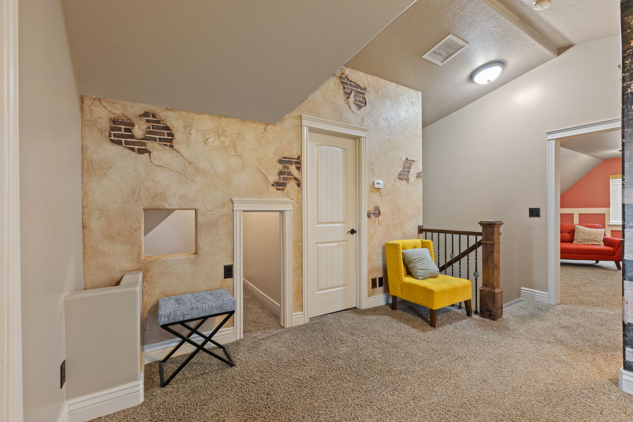 Carpeted hallway featuring a textured accent wall with exposed brick detailing, a white paneled door, and wood newel post with metal balusters