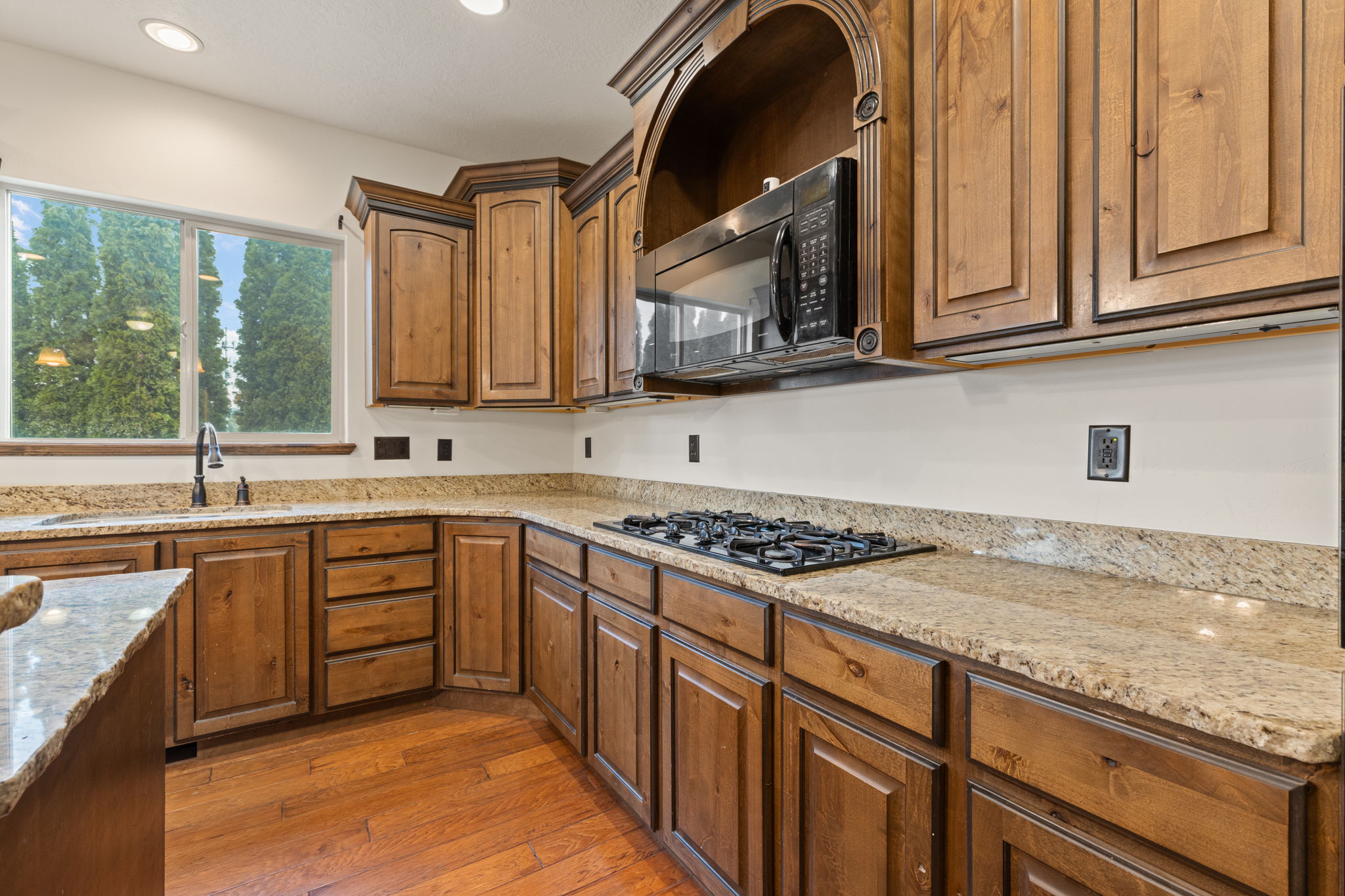 Kitchen featuring wood cabinetry, light-toned granite countertops, a built-in microwave, a gas cooktop, and wood-finish flooring
