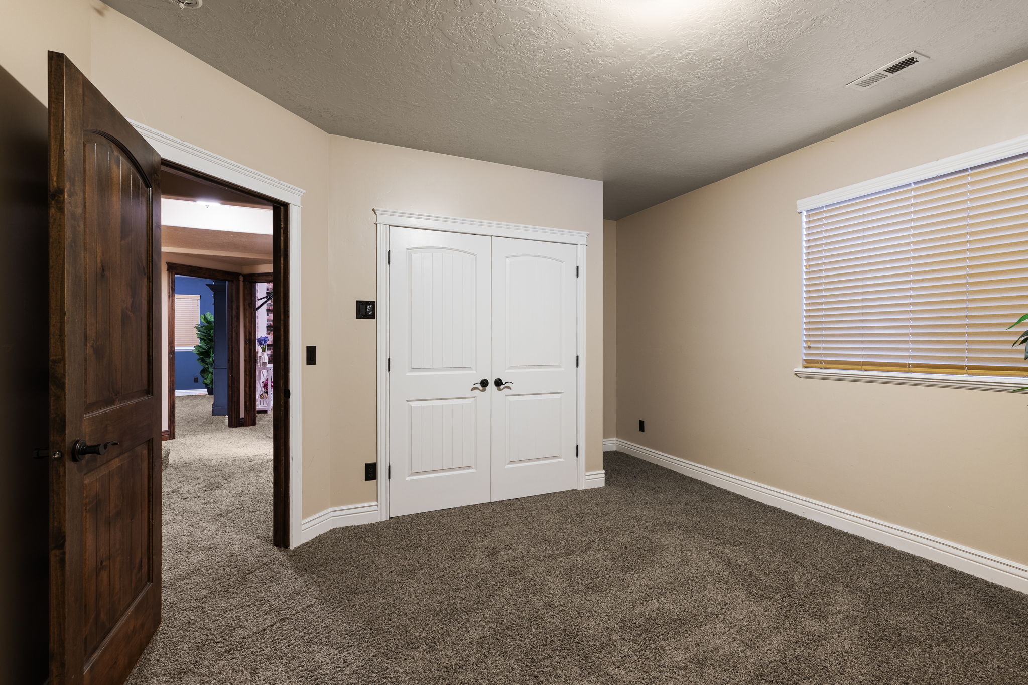 Neutral-toned room featuring a window with blinds, a double-door closet with black hardware, and textured carpet flooring