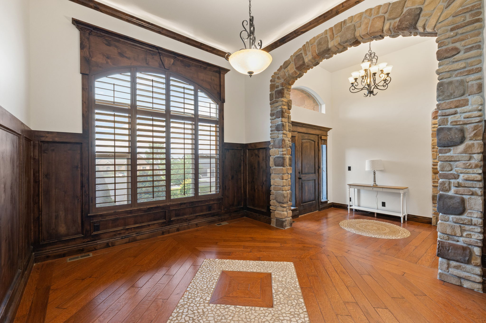 Entryway featuring stone archway, wood-finish flooring with mosaic inlay, wood paneling, and an arched window with plantation shutters