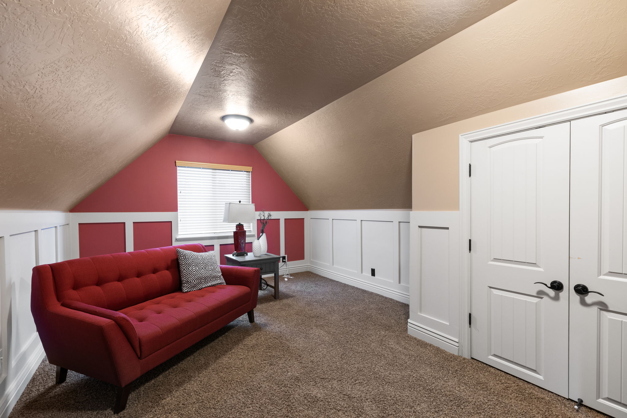 Carpeted attic space featuring white wainscoting, sloped ceilings, and a pair of white paneled doors with black hardware