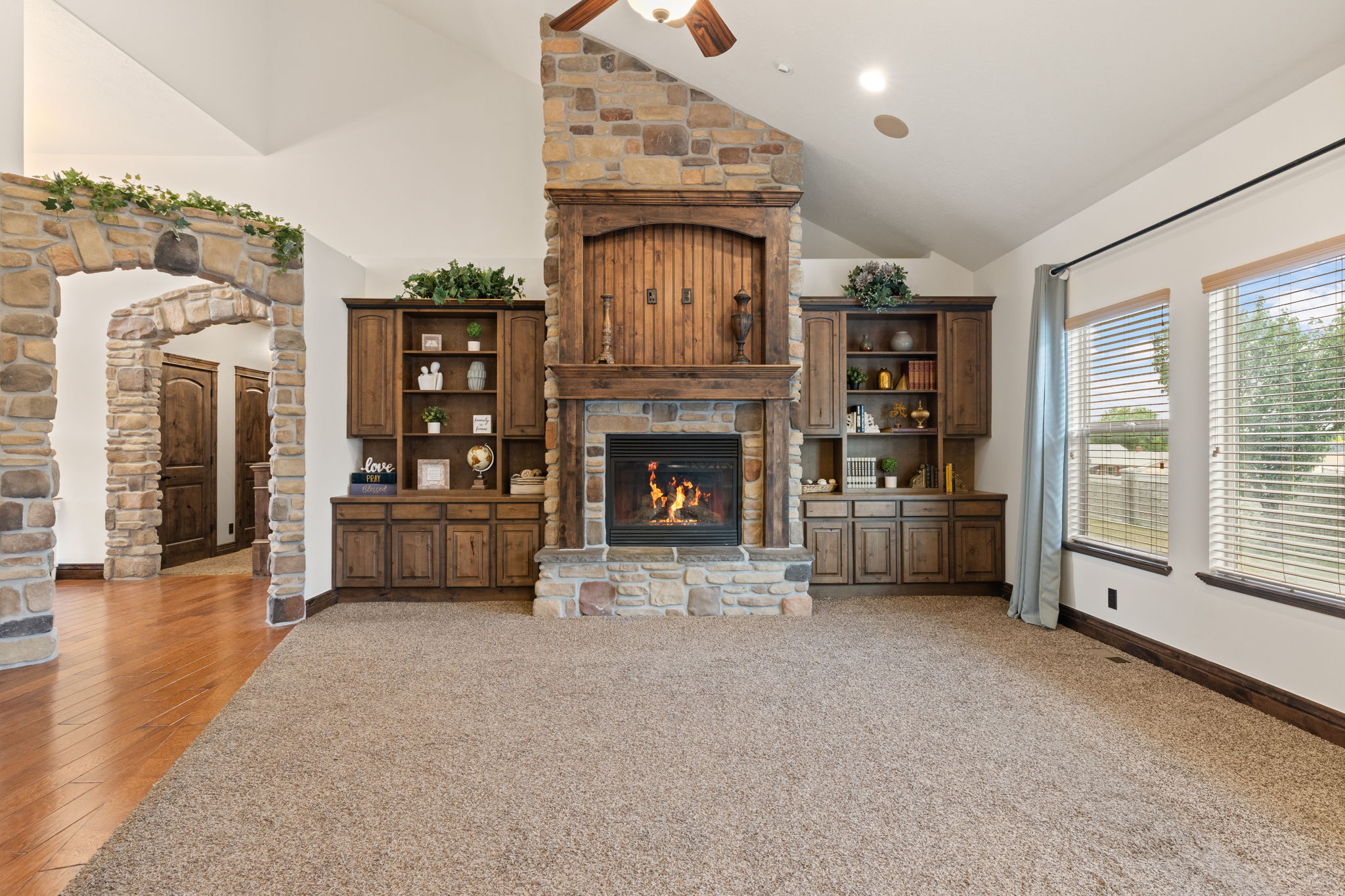 Vaulted ceiling living area featuring a stone fireplace with wood mantel, built-in cabinetry, and multiple windows with blinds