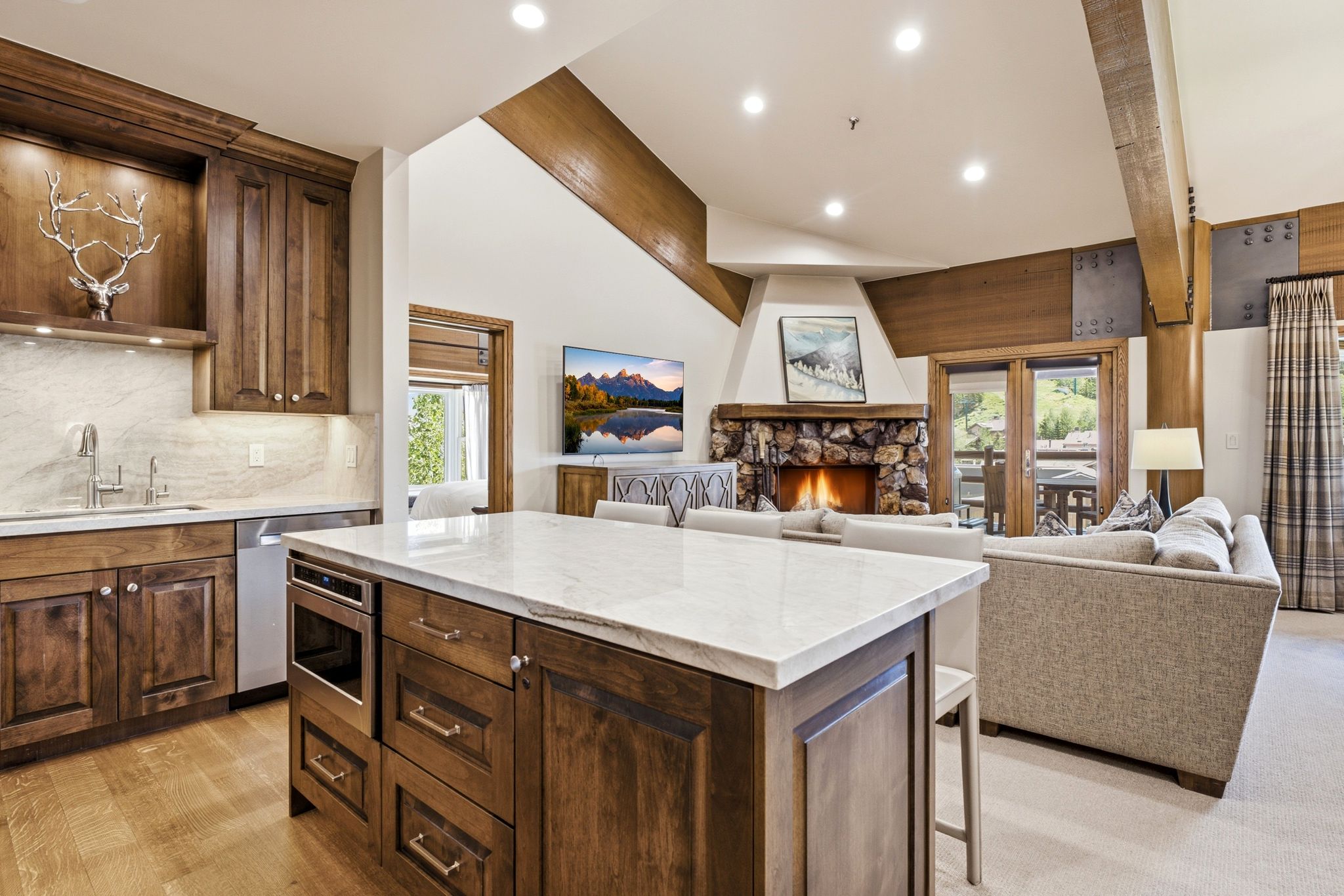 Open-concept living space featuring a stone fireplace, wood-finish flooring, exposed wood beams, a spacious kitchen island with a light-toned countertop, and dark wood cabinetry