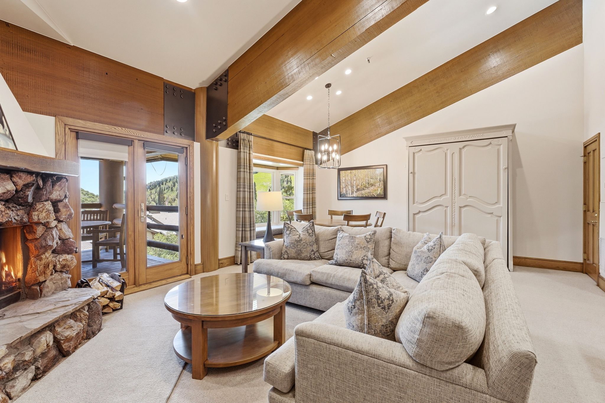 Spacious living area featuring a floor-to-ceiling stone fireplace, vaulted ceiling with wood beams, and direct access to an outdoor balcony