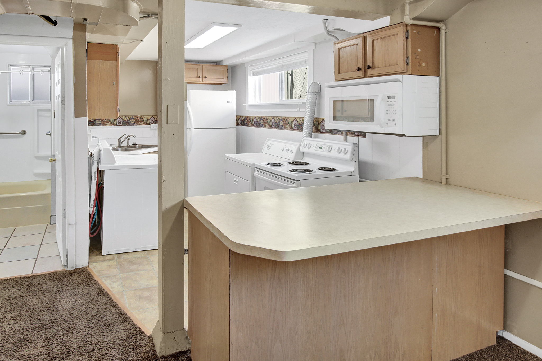 Kitchenette featuring light wood-finish cabinetry, white appliances, and a light-toned countertop with an integrated breakfast bar