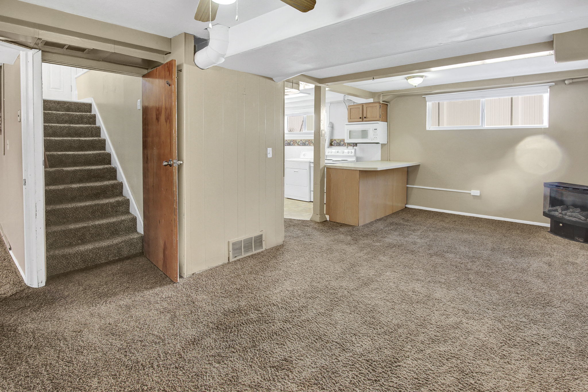 Carpeted open-concept lower level featuring a built-in breakfast bar with a light-toned countertop, a kitchen area with upper and lower cabinetry, and a window providing ambient light