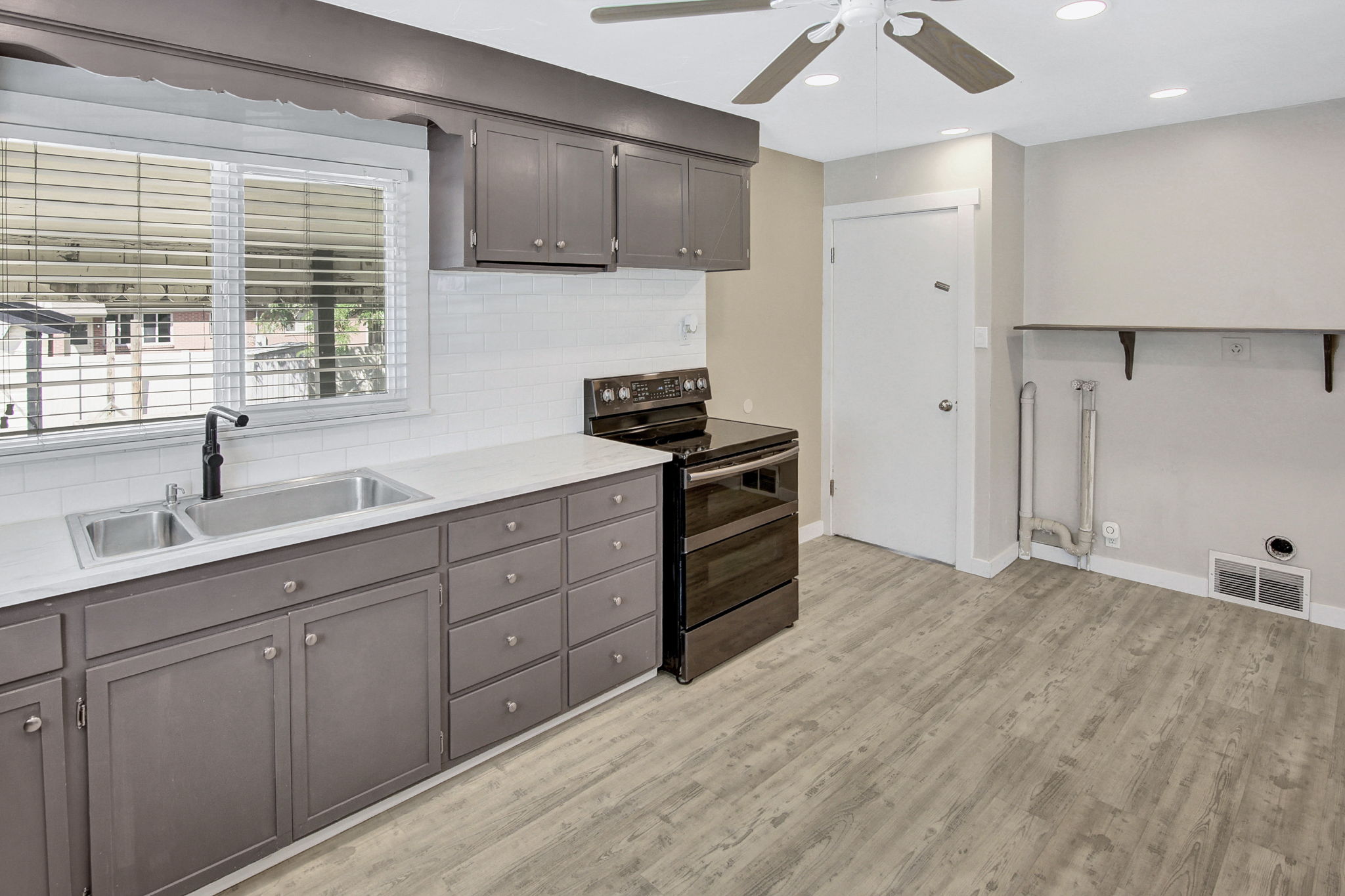 Kitchen area featuring dark gray cabinetry, a white subway tile backsplash, a stainless steel double basin sink with a matte black faucet, and light wood-finish flooring