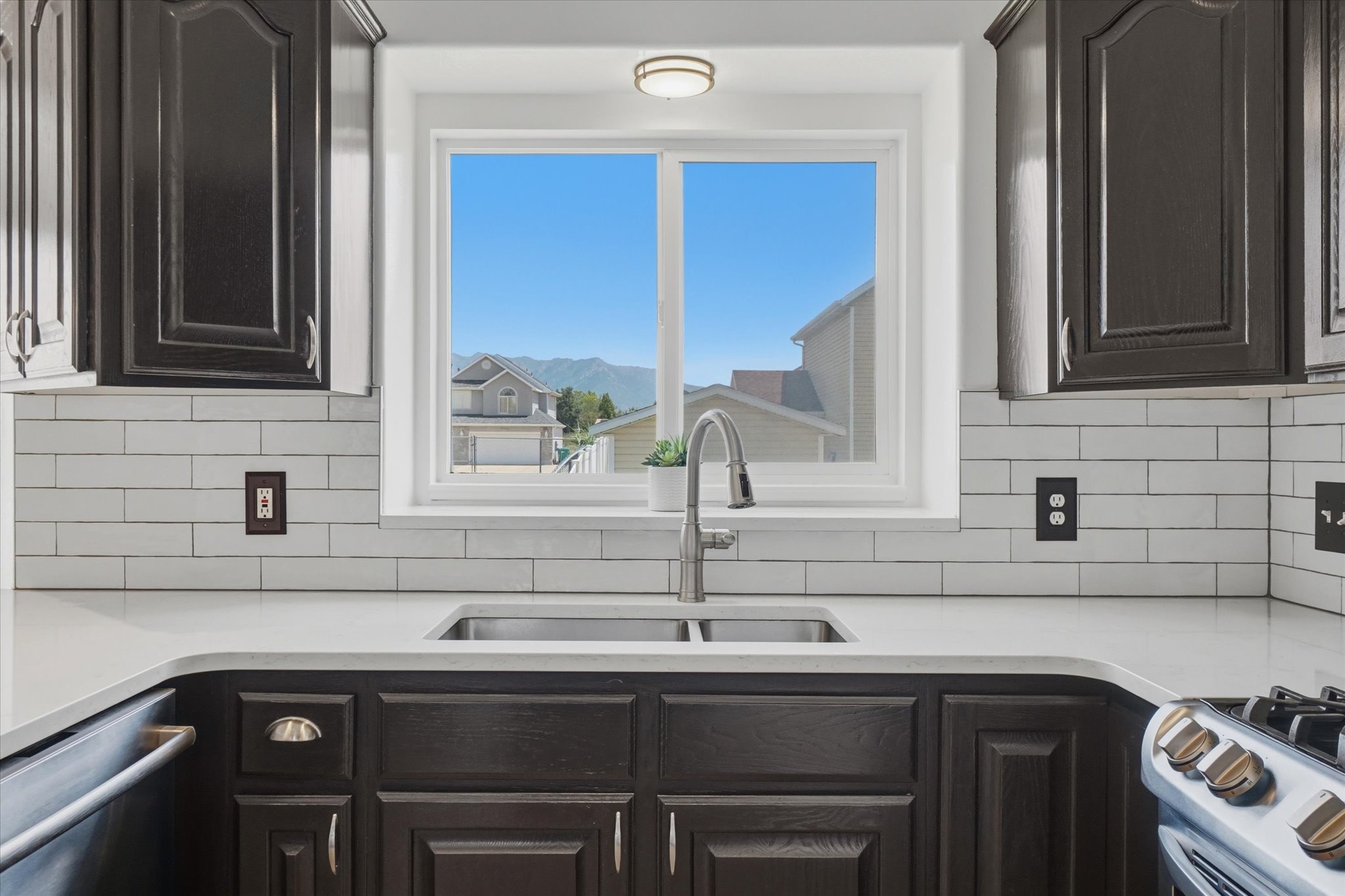 Kitchen sink area featuring a dual basin stainless steel sink with a gooseneck faucet, light-toned countertops, subway tile backsplash, dark wood cabinetry, and a window with a mountain backdrop
