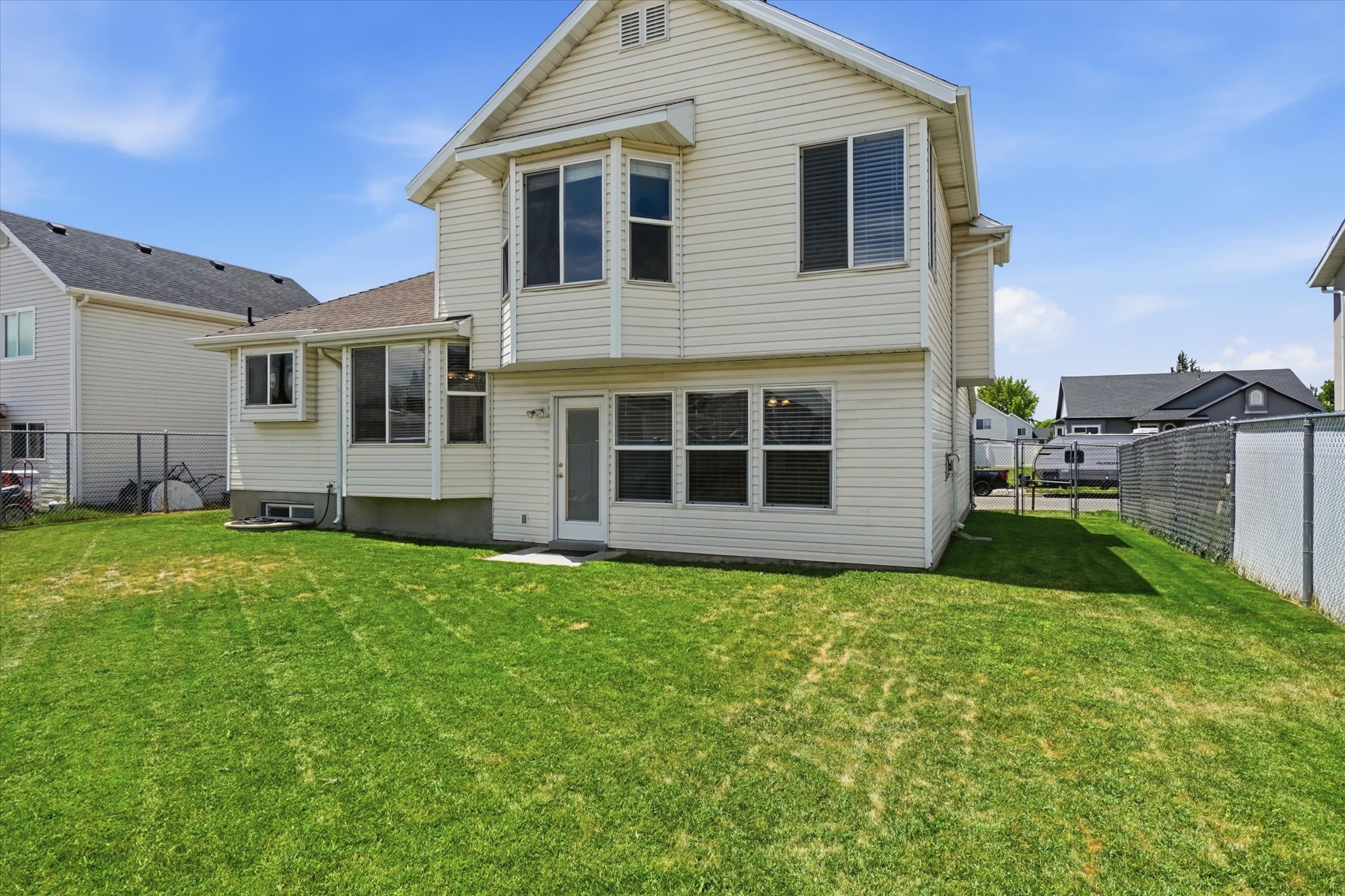 Expansive rear yard featuring a verdant lawn, a white vinyl-sided exterior, a chain-link fence, multiple windows with blinds, and a rear entry door