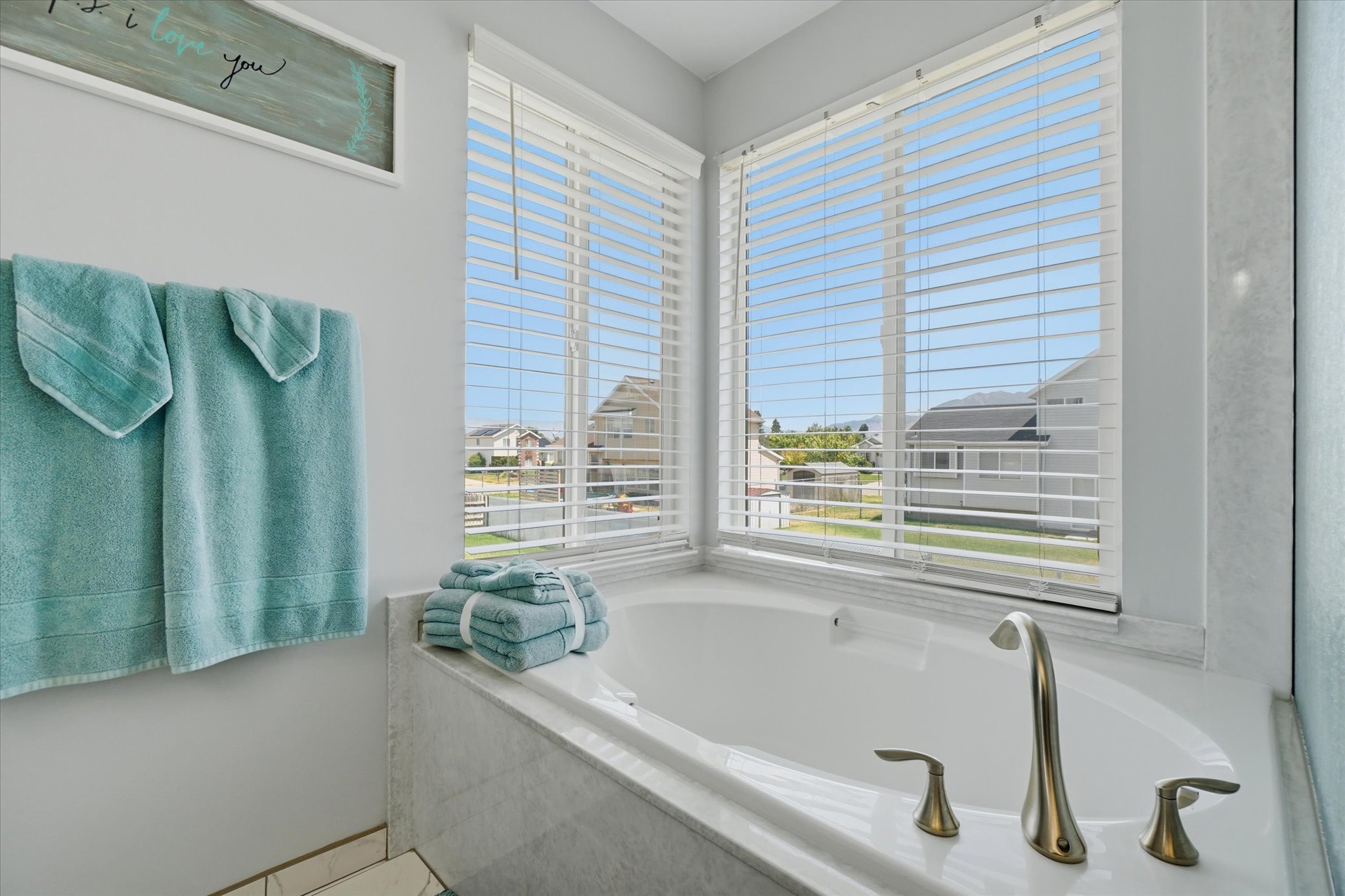 Soaking tub surrounded by light-toned tile, featuring brushed nickel fixtures and two large windows with horizontal blinds