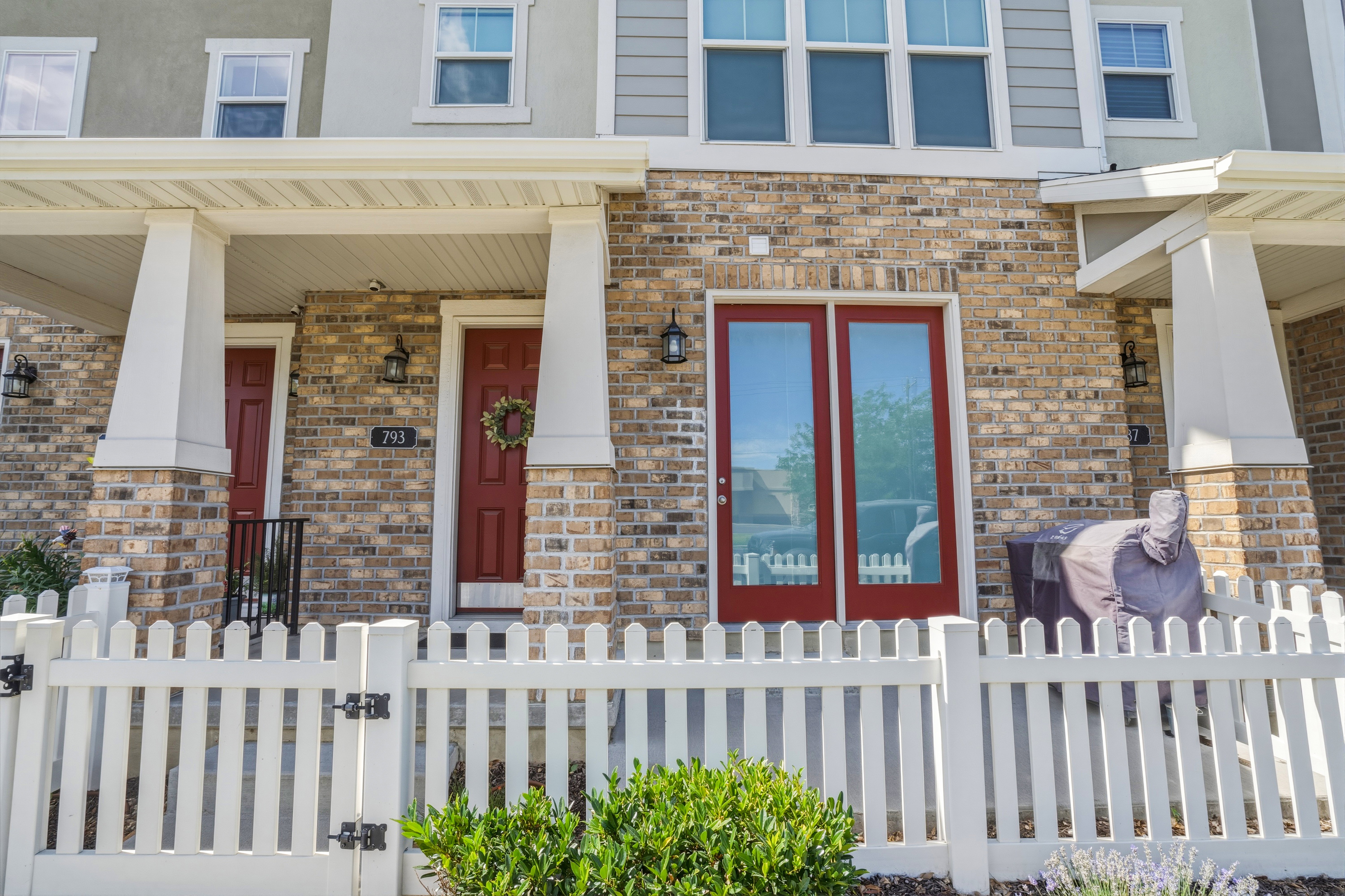 Brick facade townhome with a white picket fence, covered front porch, red entry door, and red-framed glass patio doors