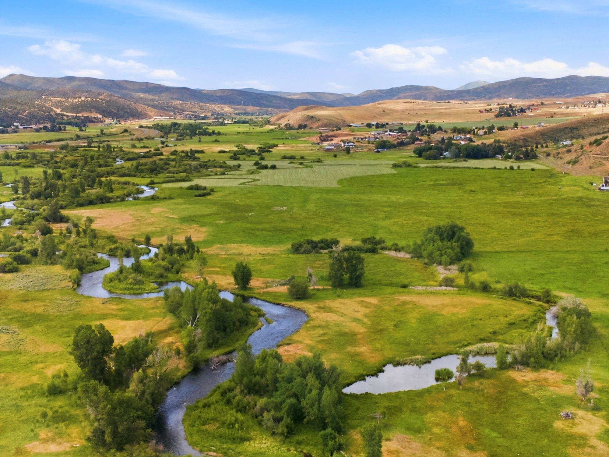 Expansive green landscape featuring a winding river, mature trees, distant hills, and clear skies