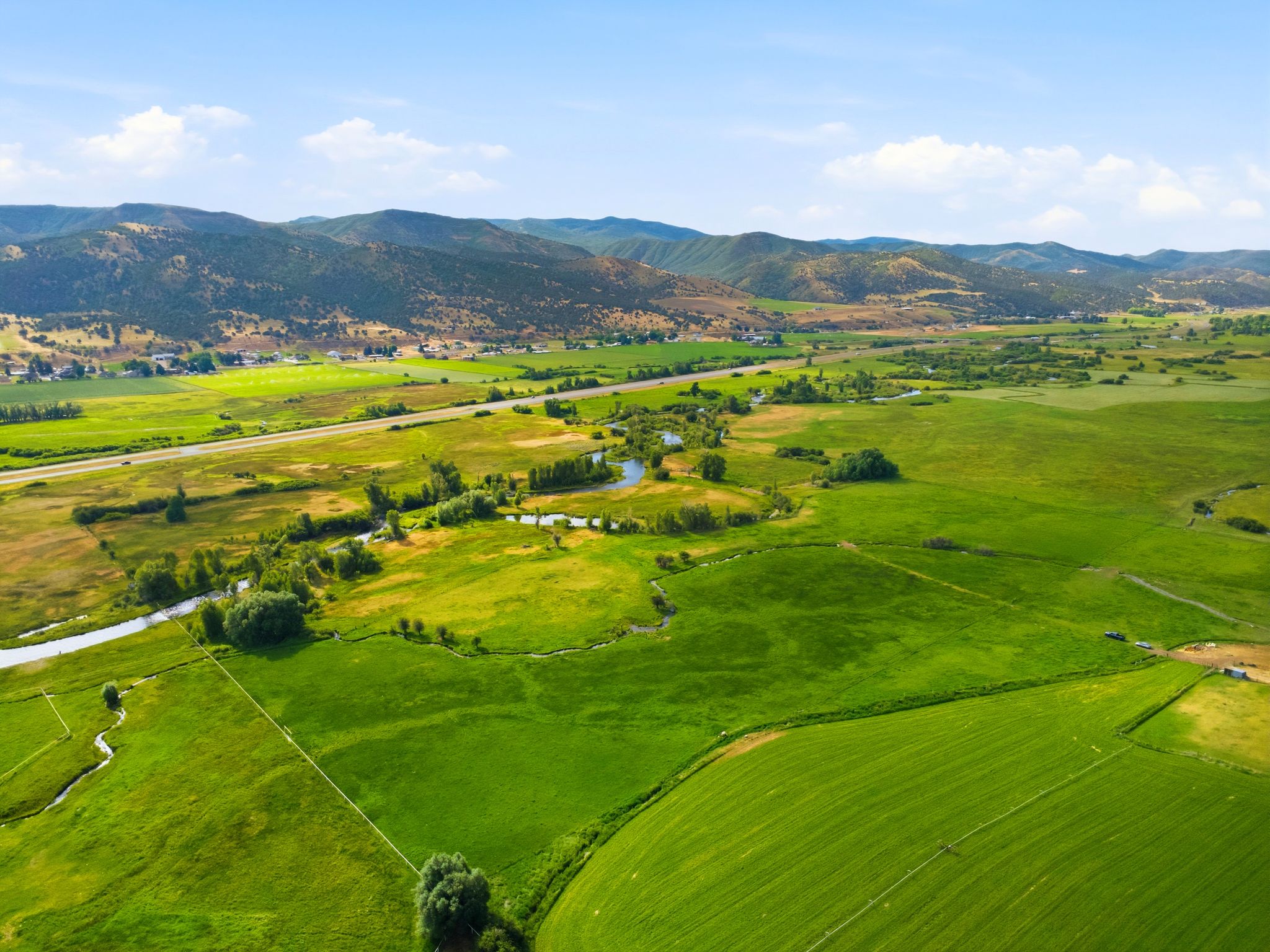 Expansive green fields featuring natural waterways, a paved roadway, and distant mountain ranges under a clear sky
