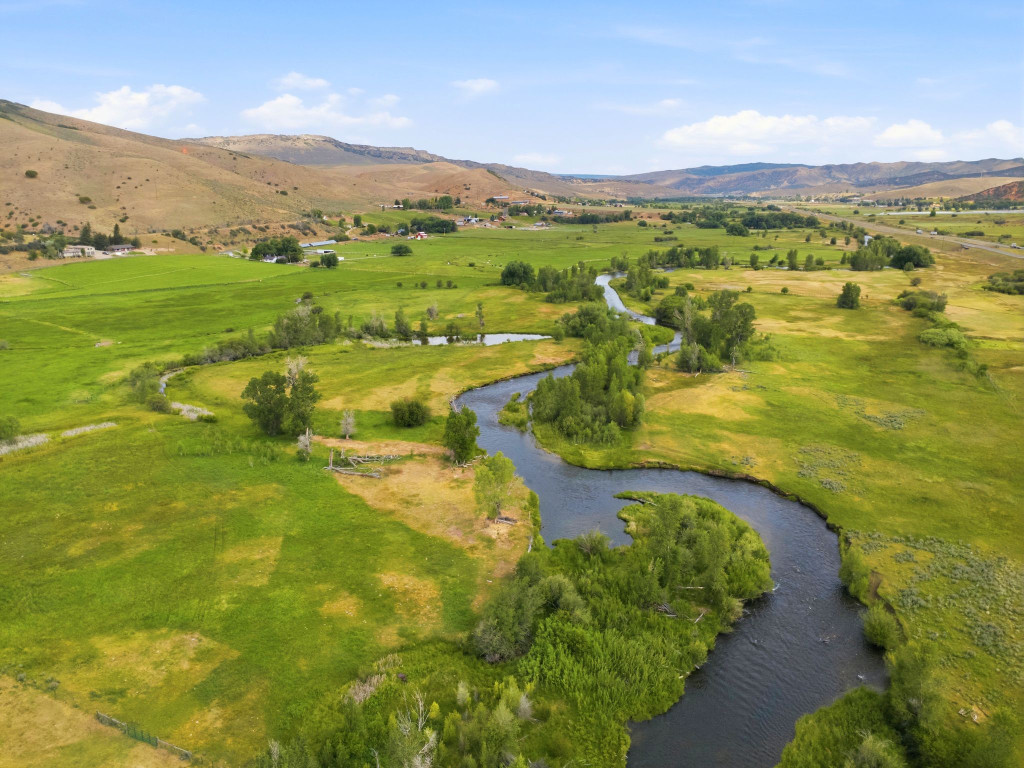 Expansive natural landscape featuring a meandering river, lush green fields, scattered mature trees, and distant rolling hills under a clear sky